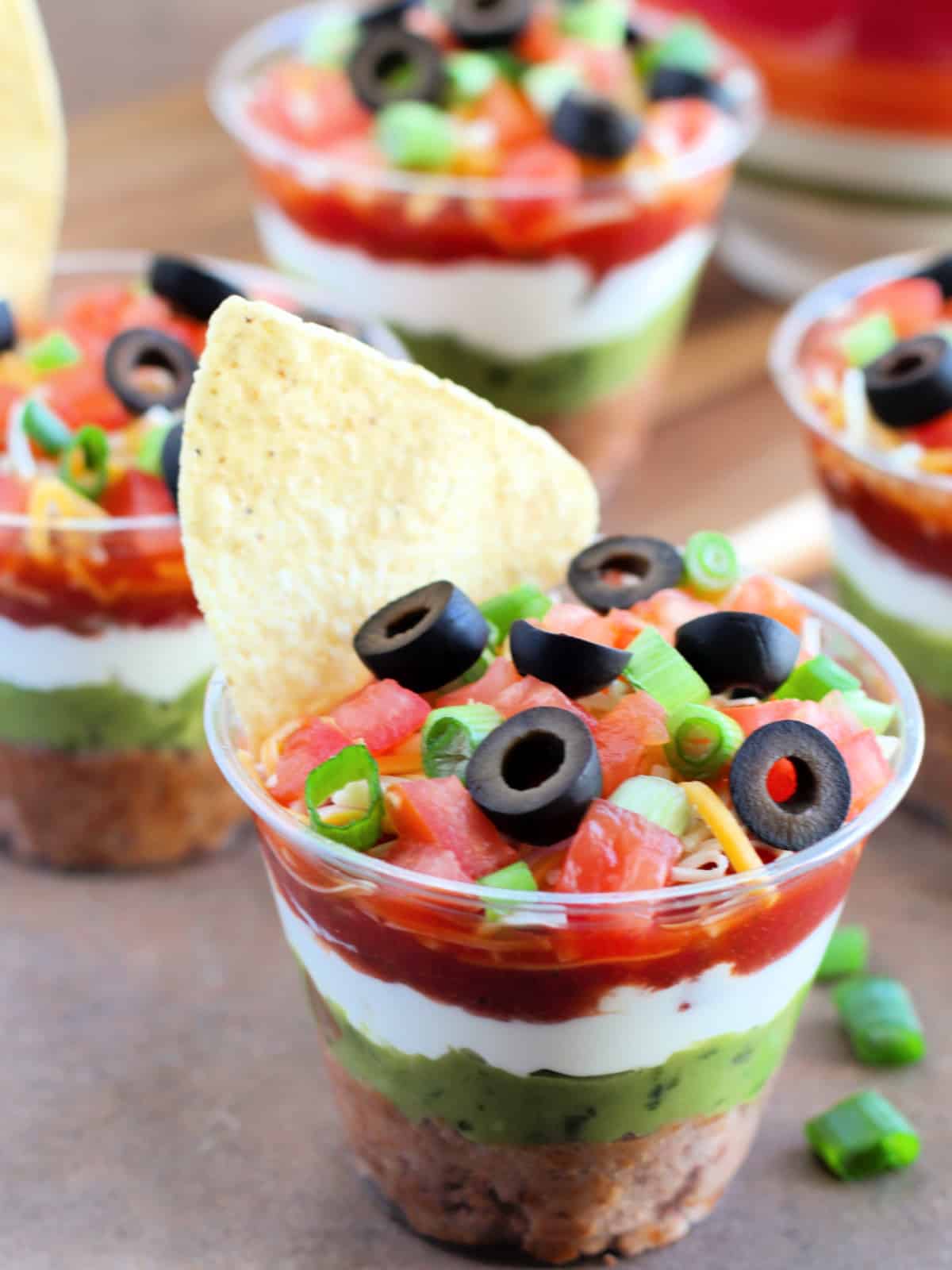 Clear cups filled with layered dip, topped with sliced olives and chopped green onions, with tortilla chips inserted on top.