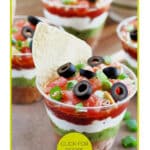 Clear cups filled with layered dip, topped with black olives, tomatoes, and green onions. A tortilla chip is inserted into each cup. Text reads "7-Layer Dip Cups" and "Click for Recipe Ideas.