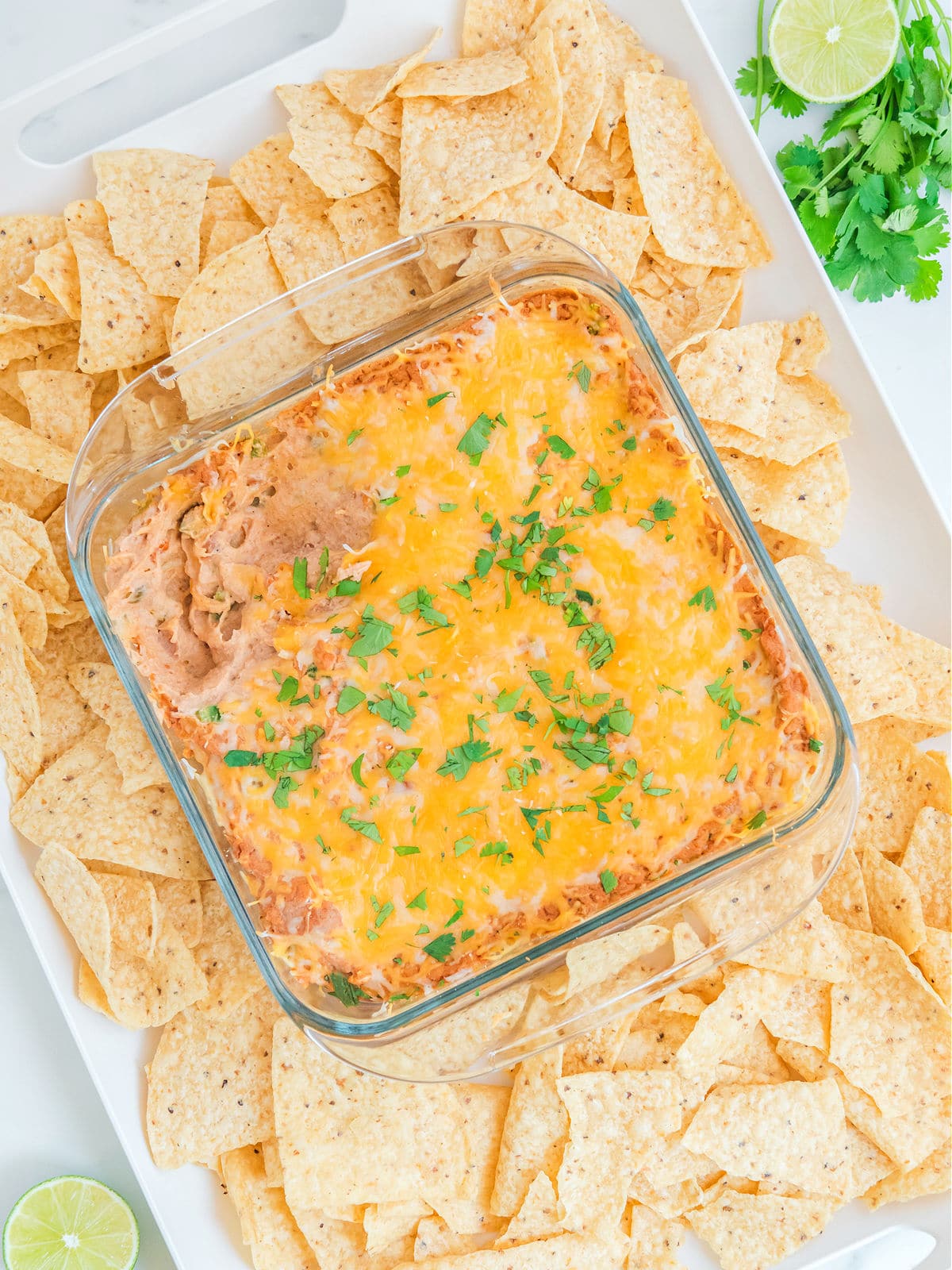 Square glass dish of cheesy bean dip garnished with herbs, surrounded by tortilla chips on a tray. Lime and cilantro are placed in the corner.