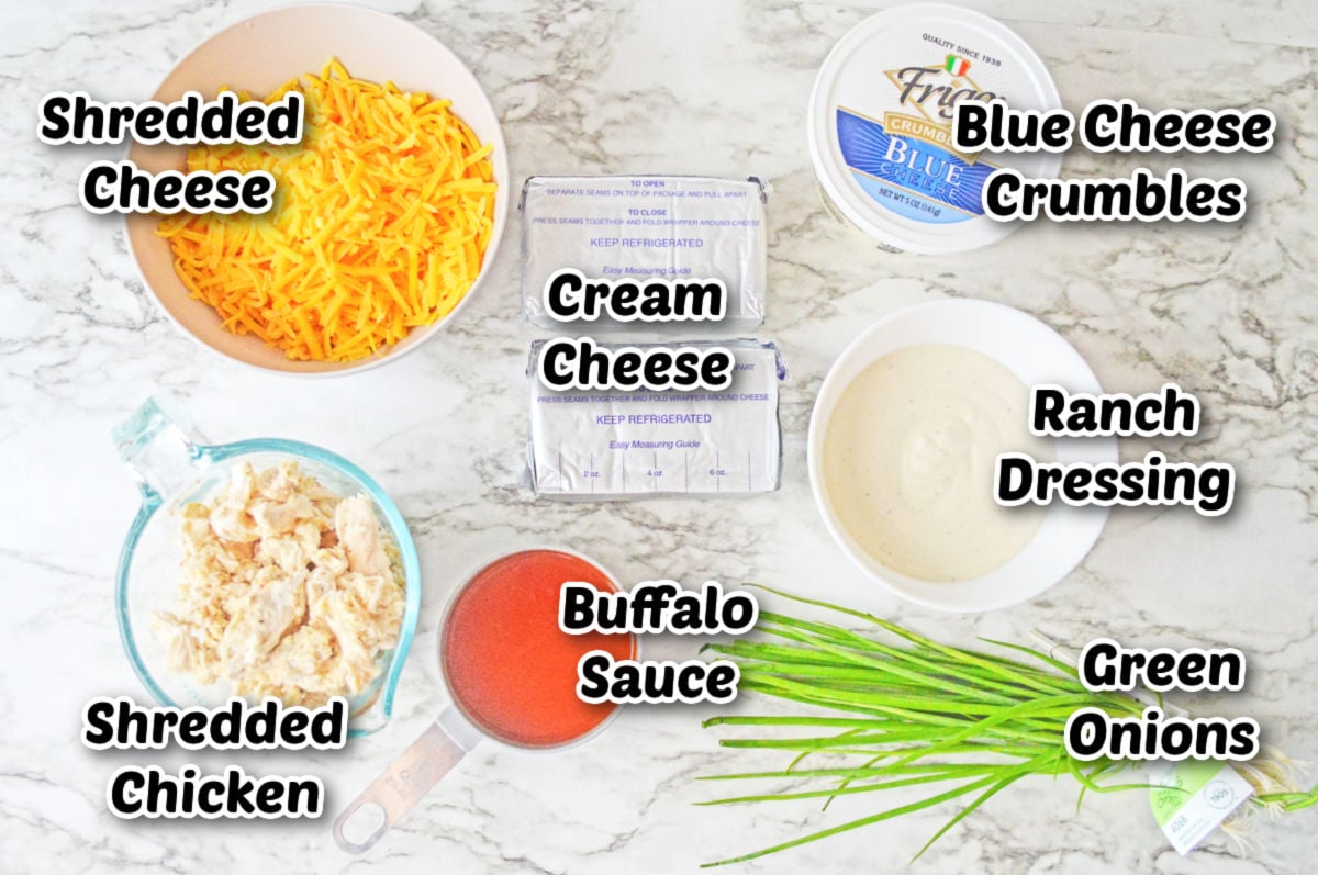 Ingredients for buffalo chicken dip on a marble surface: shredded cheese, cream cheese, blue cheese crumbles, ranch dressing, shredded chicken, buffalo sauce, and green onions.