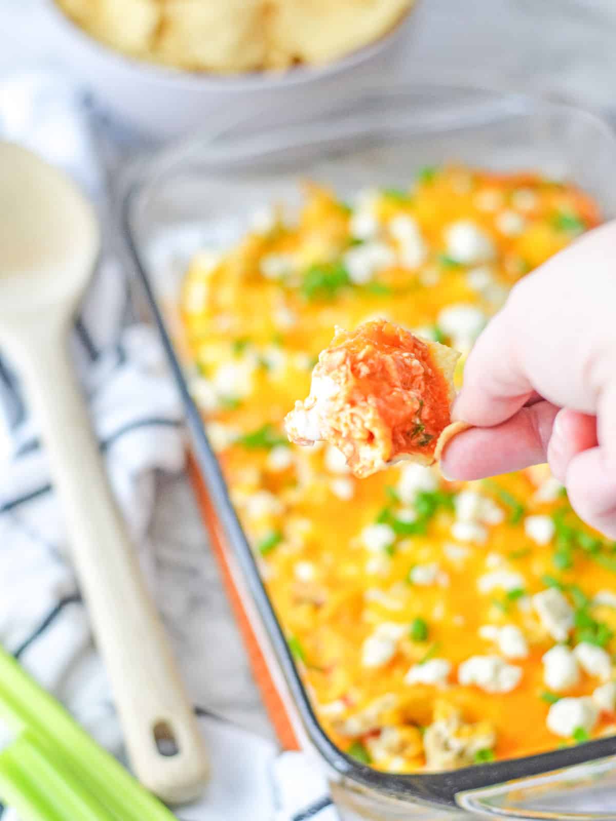 A hand holding a chip with buffalo chicken dip over a glass baking dish filled with melted cheese dip, garnished with herbs. A wooden spoon and green onions are nearby.