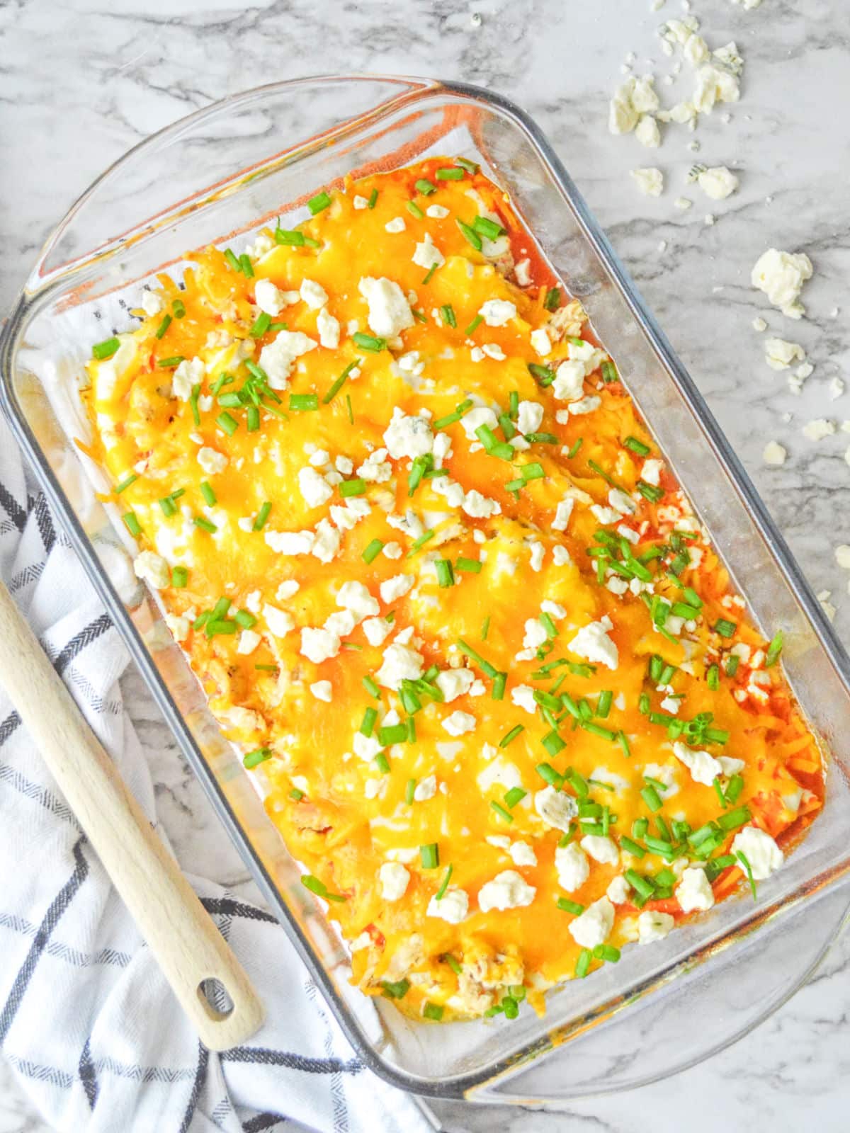 Baked casserole in a glass dish with melted cheese, green onions, and crumbled feta, on a marble surface next to a striped towel.