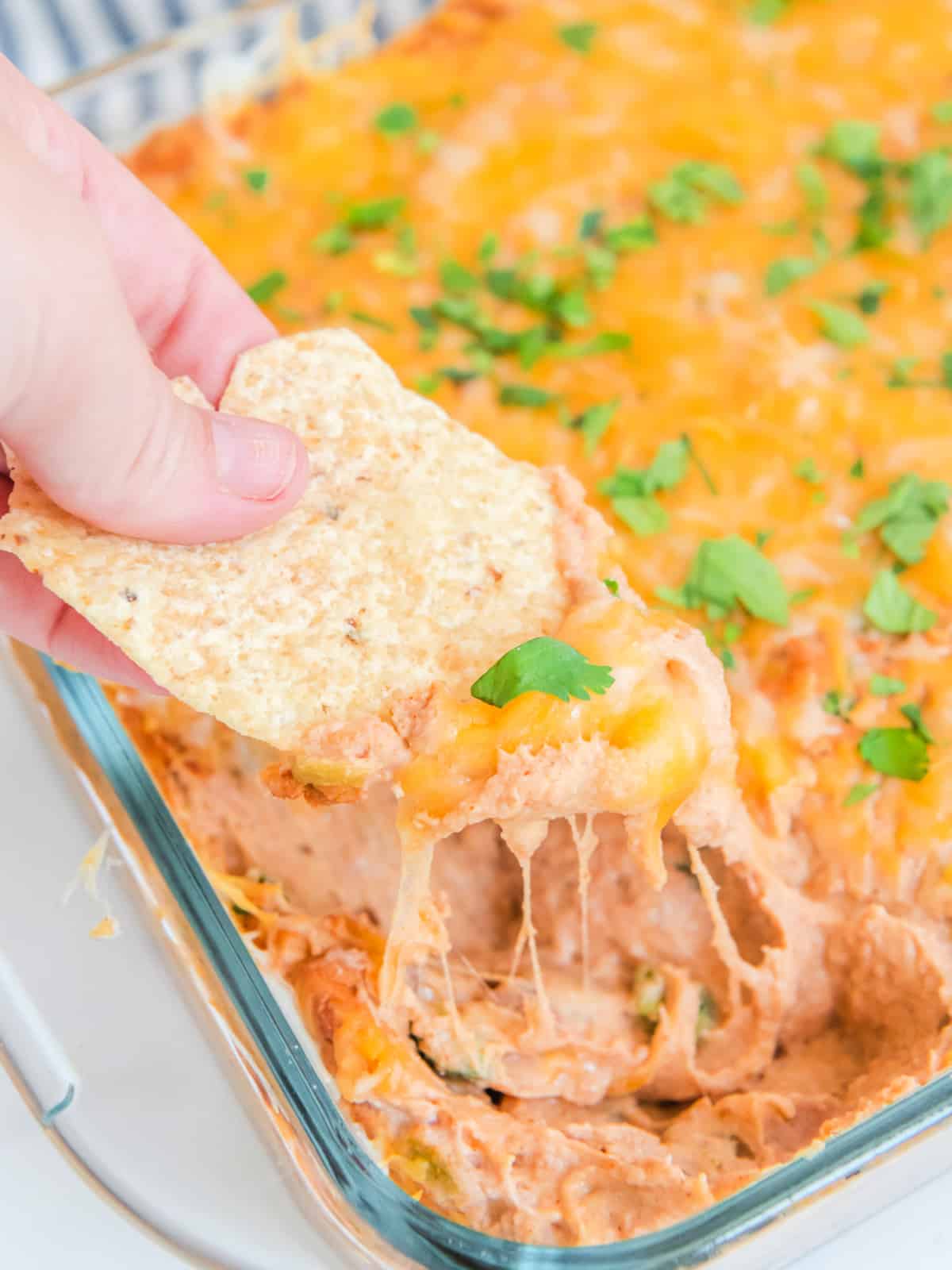 A hand dips a tortilla chip into a cheesy bean dip in a glass baking dish, garnished with chopped green herbs.