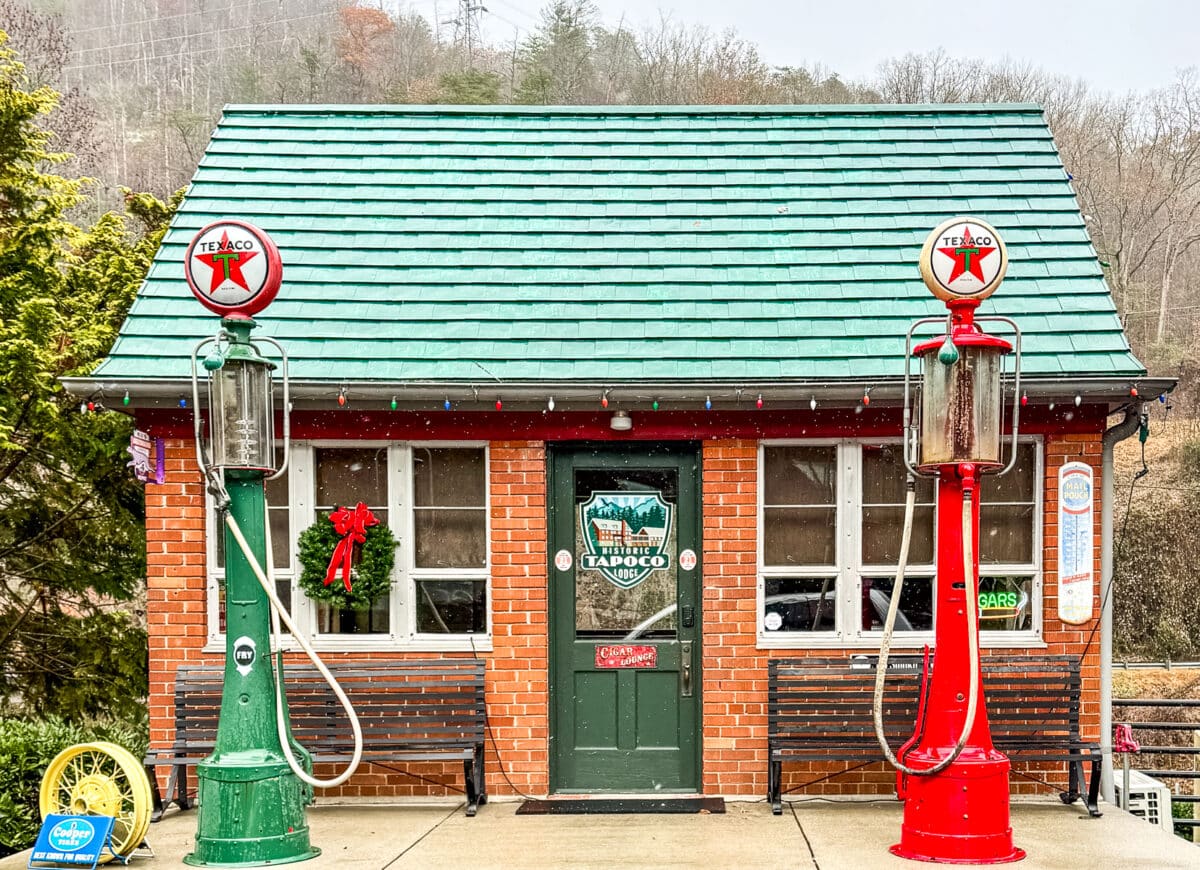 A small brick building with a green roof, flanked by vintage Texaco gas pumps, one green and one red. It has a wreath on the door and a sign in the window.