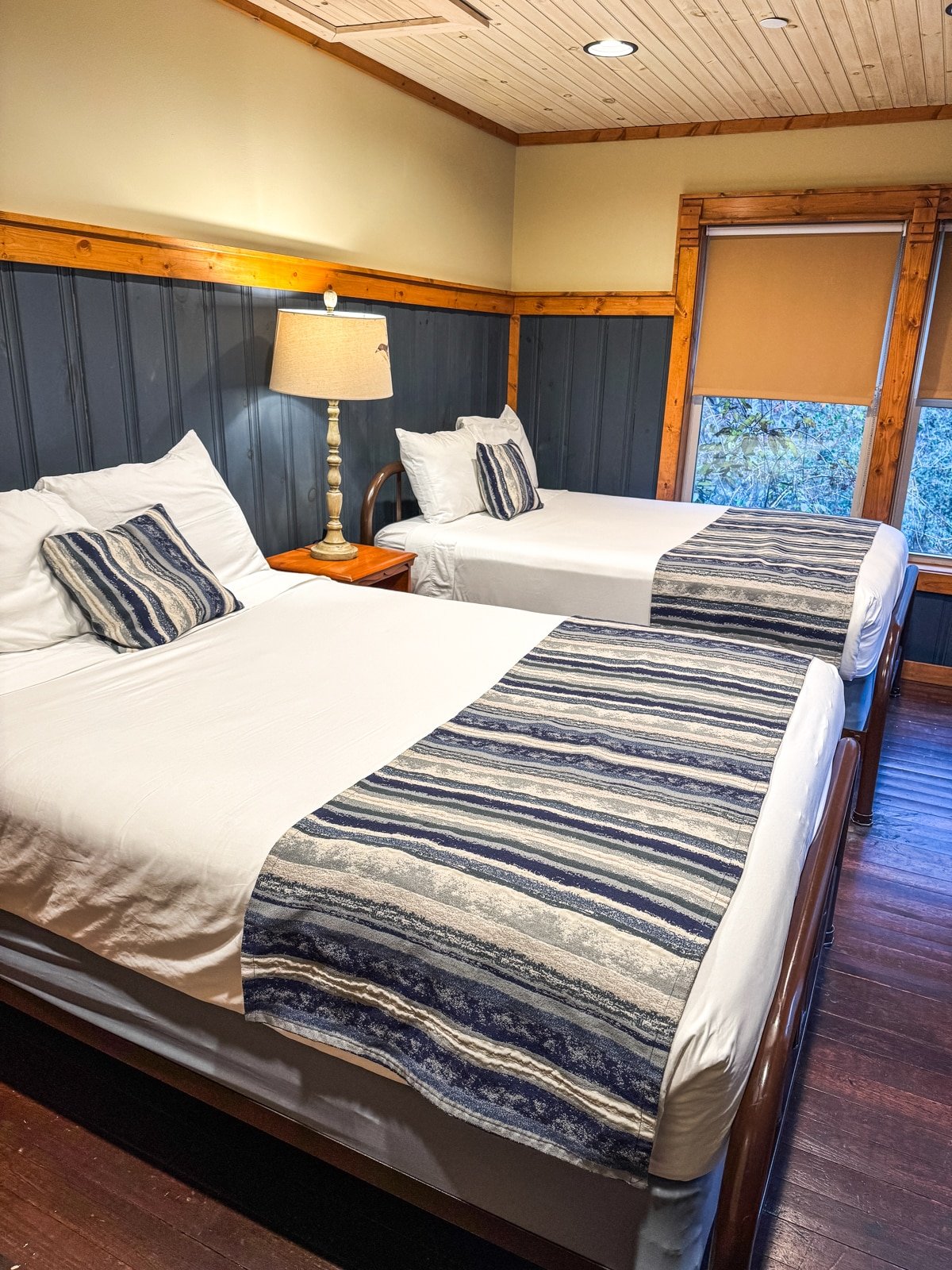 A cozy bedroom with two neatly made beds, striped blue and white bed runners, a shared bedside table with a lamp, and a window covered by a shade.