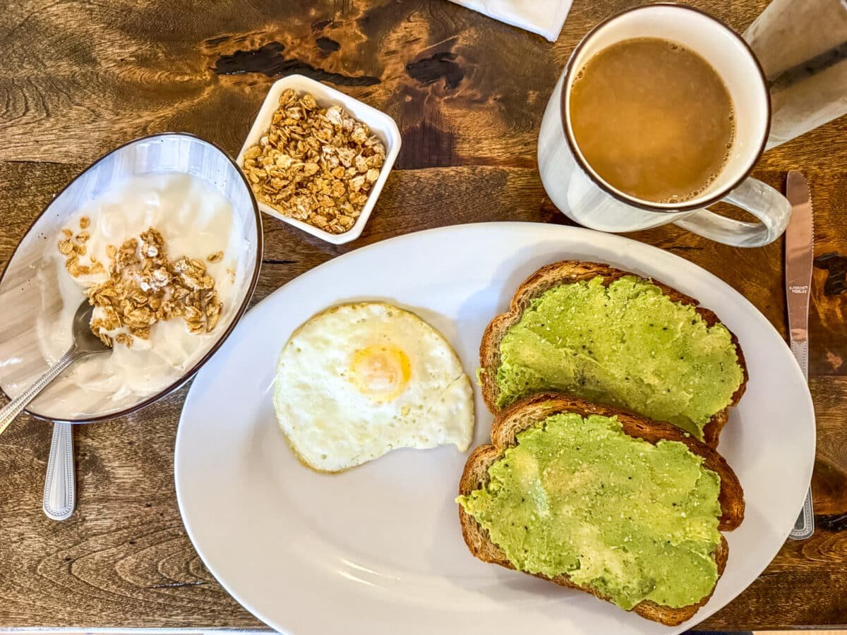 Breakfast of avocado toast, a fried egg, yogurt with granola, and a cup of coffee on a wooden table.