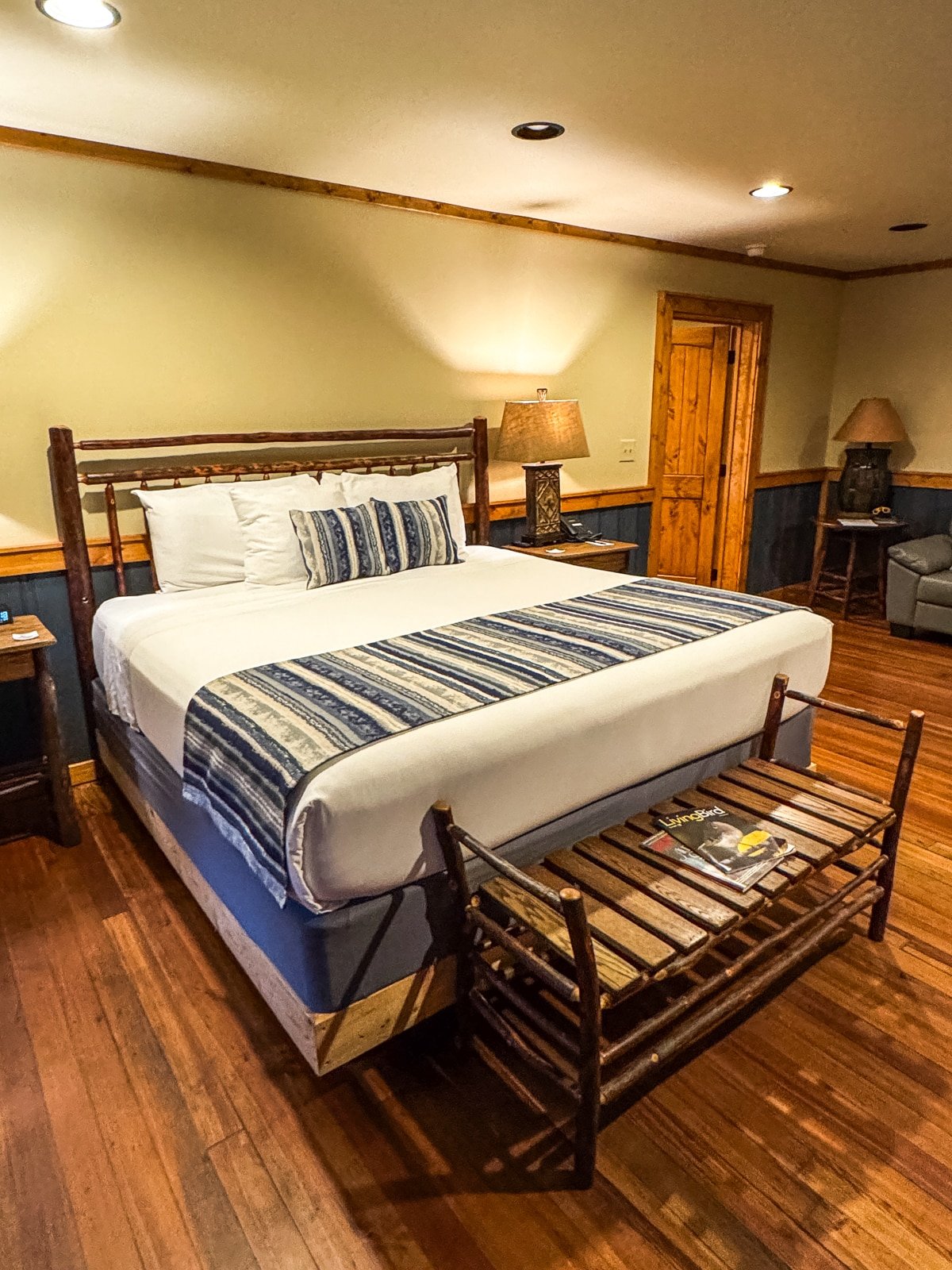 A rustic bedroom with a wooden bed, blue and white striped bedding, wooden bench at the foot, nightstands with lamps, and wooden flooring.