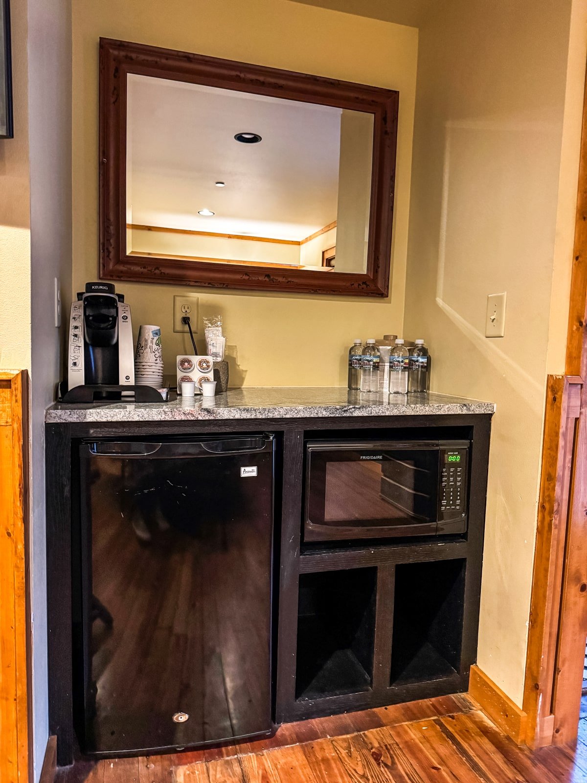 A small kitchenette with a mirror above, featuring a coffee maker, cups, bottles of water, a mini fridge, and a microwave on a granite countertop.