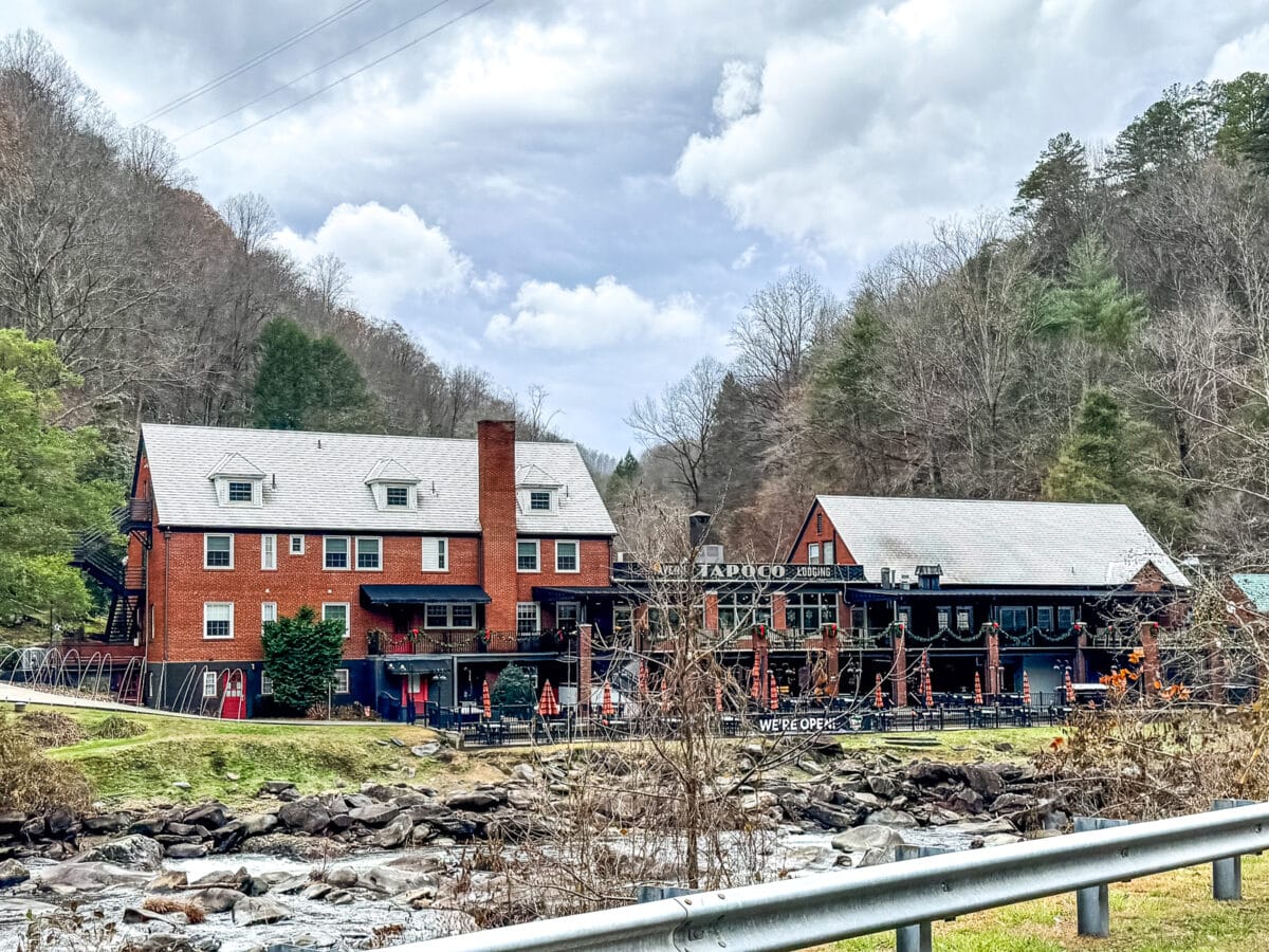 A brick building and a wooden structure stand beside a rocky creek, surrounded by trees under a cloudy sky.