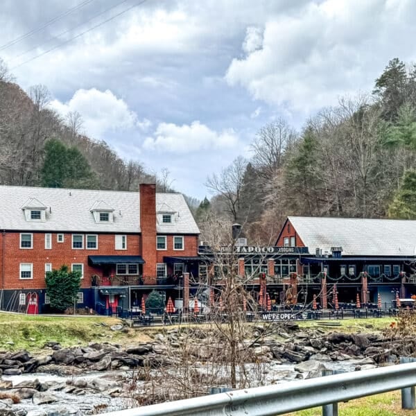 A brick building and a wooden structure stand beside a rocky creek, surrounded by trees under a cloudy sky.