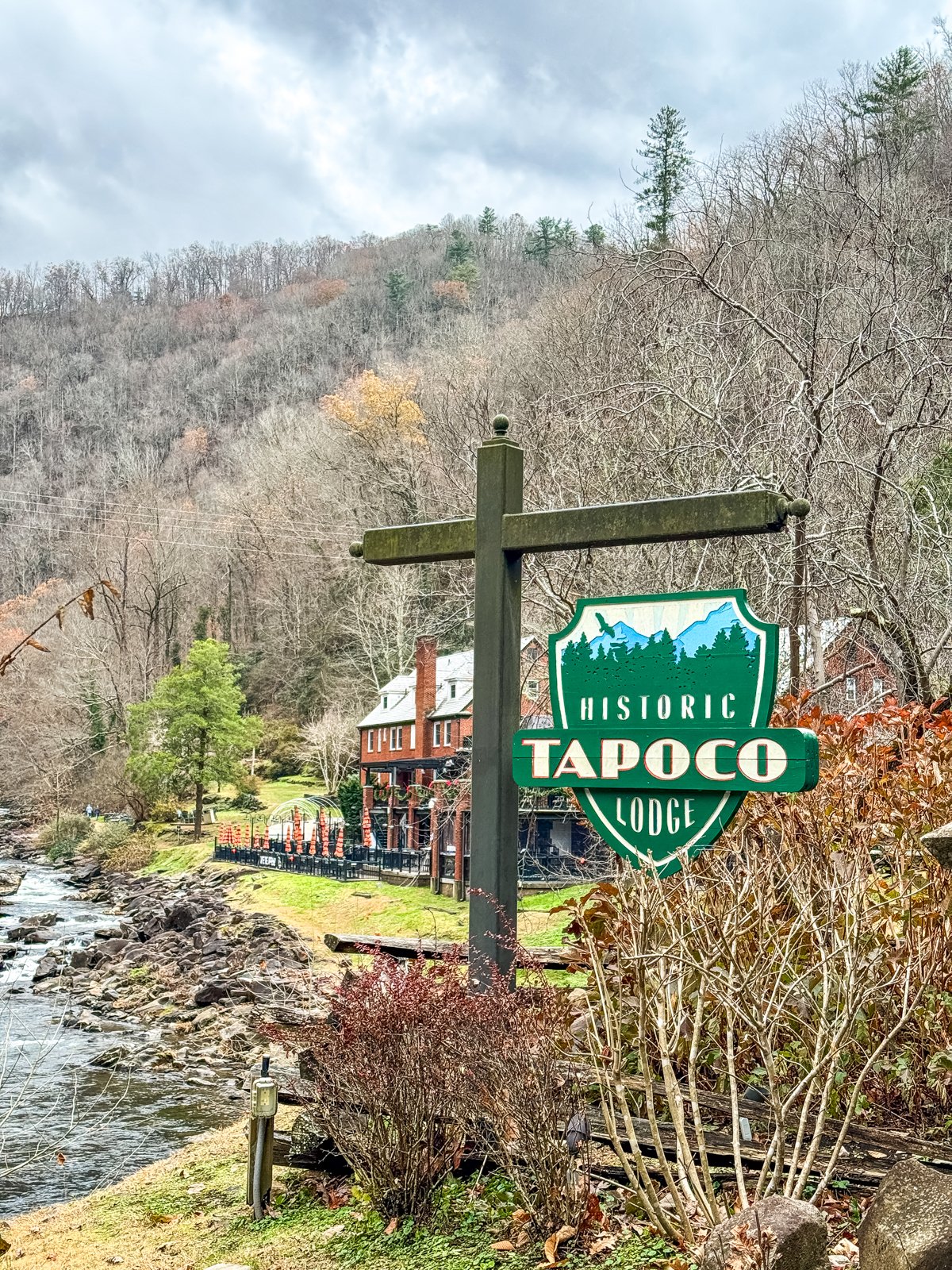 Sign for Historic Tapoco Lodge in a wooded area near a river, with lodge buildings in the background. Overcast sky.