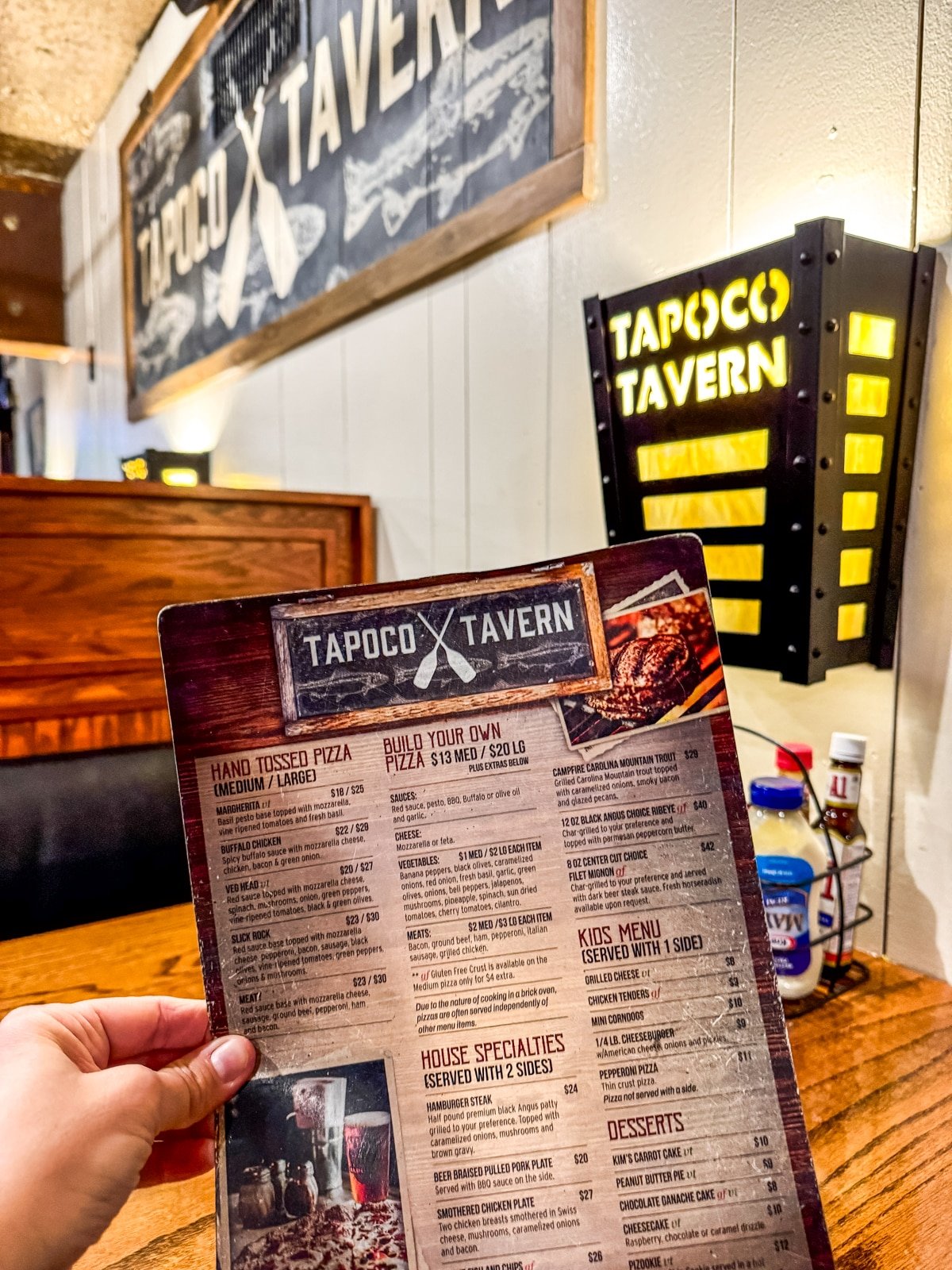 A person holds a Tapoco Tavern menu inside the restaurant. The interior features wood accents and wall signs.