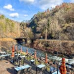 Riverside outdoor dining area with empty tables and umbrellas. A peaceful river flows alongside, flanked by a forested hillside. A bridge crosses the river in the background under a blue sky.