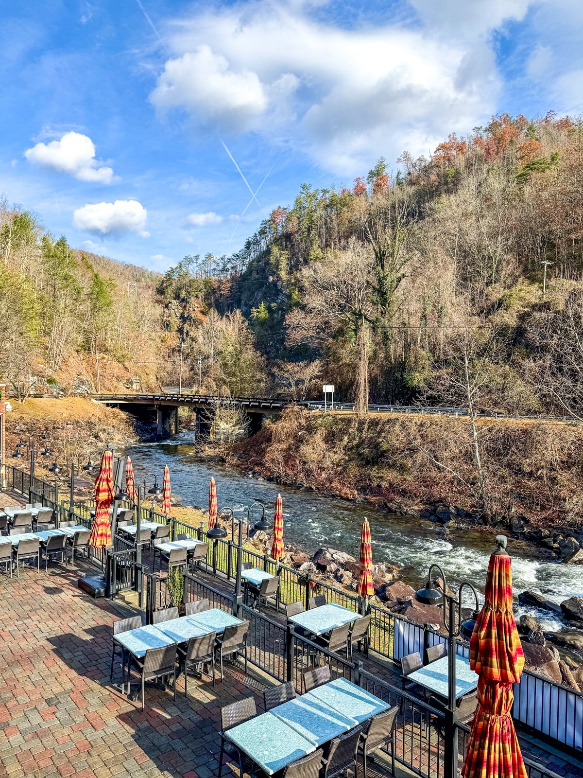 Riverside outdoor dining area with empty tables and umbrellas. A peaceful river flows alongside, flanked by a forested hillside. A bridge crosses the river in the background under a blue sky.