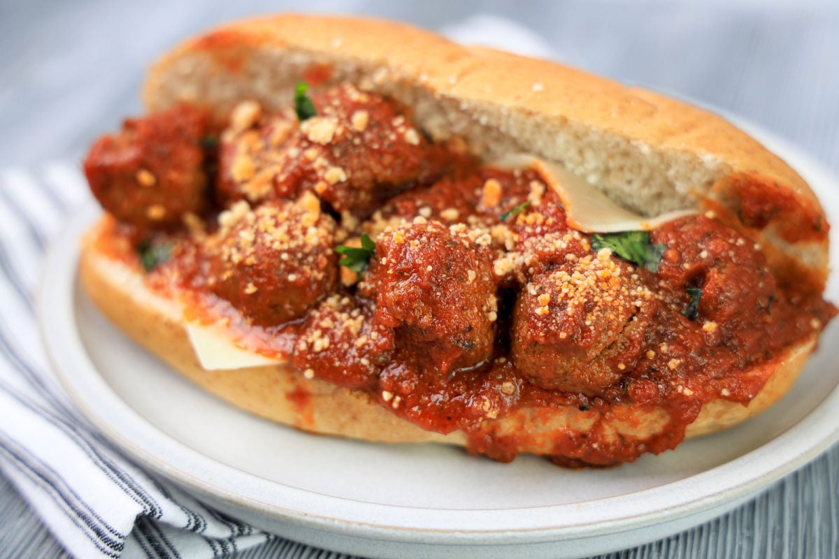 Meatball sub on a plate with tomato sauce, cheese, and sprinkled herbs, placed on a striped napkin.