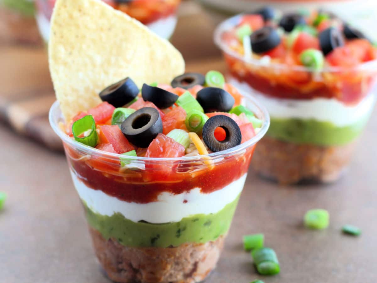 A layered dip in a clear cup features refried beans, guacamole, sour cream, salsa, diced tomatoes, sliced black olives, and green onions, topped with a tortilla chip.