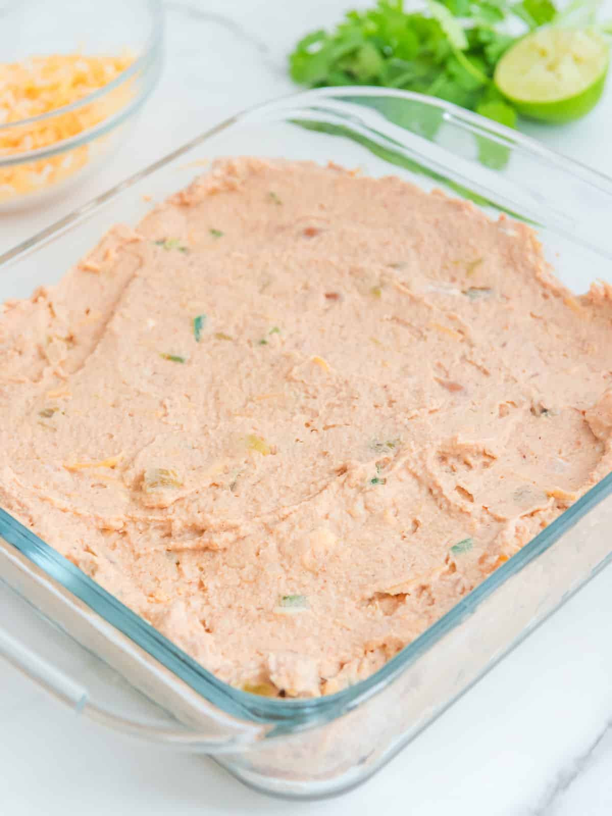 A glass square dish filled with a light orange bean dip with herbs, on a marble surface. A bowl of cheese, chopped cilantro, and a sliced lime are in the background.