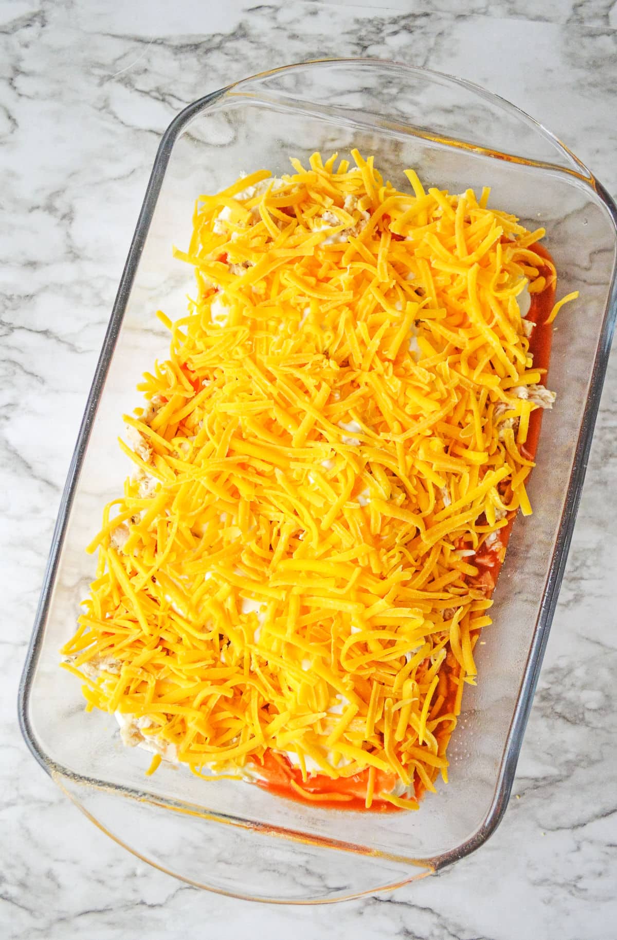 Glass baking dish filled with layers of shredded cheese over a visible layer of sauce, placed on a marble countertop.