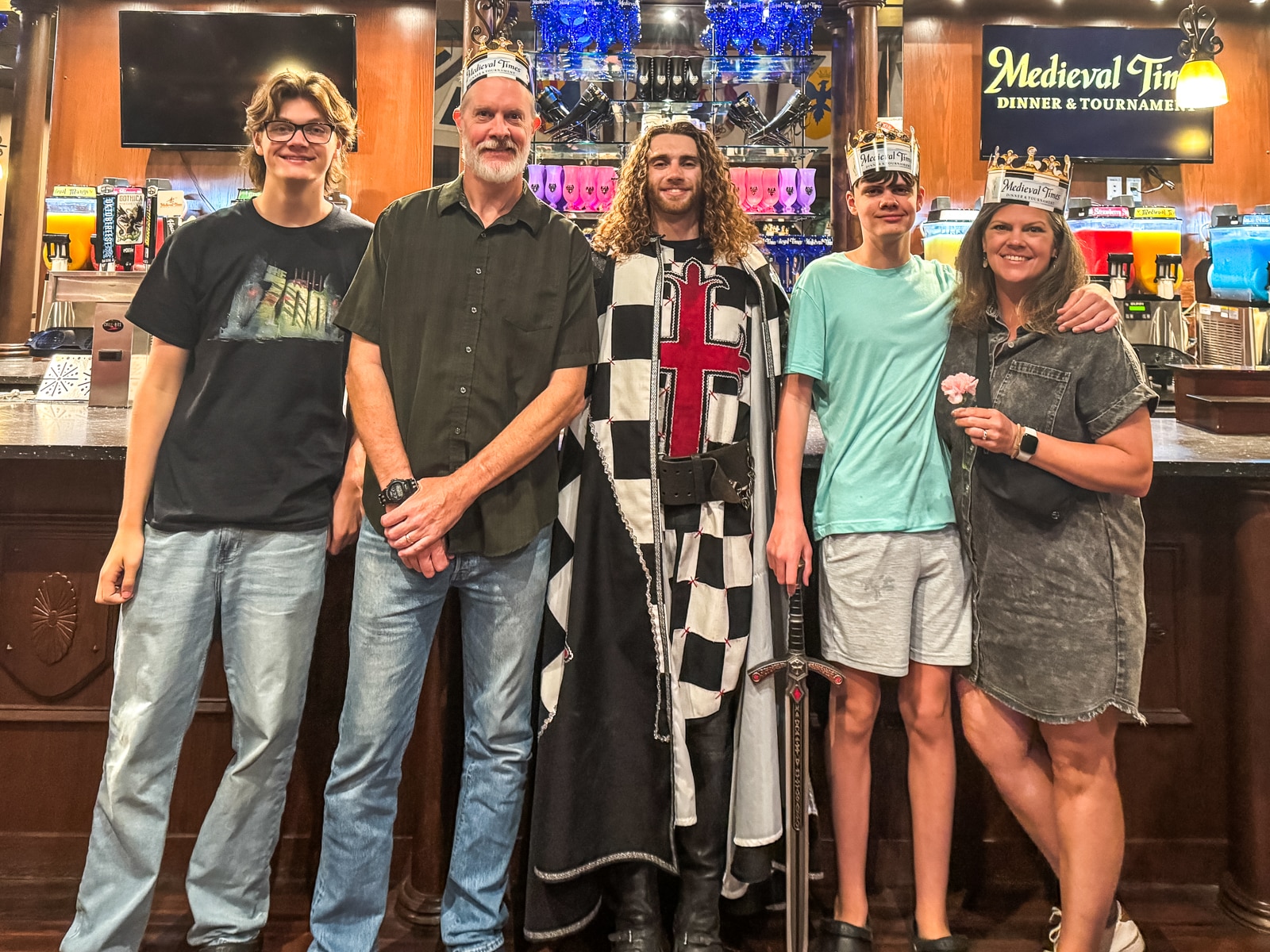 Five people stand in front of a Medieval Times sign; one is dressed as a knight, and two wear crowns. They are posing together in a casual setting.
