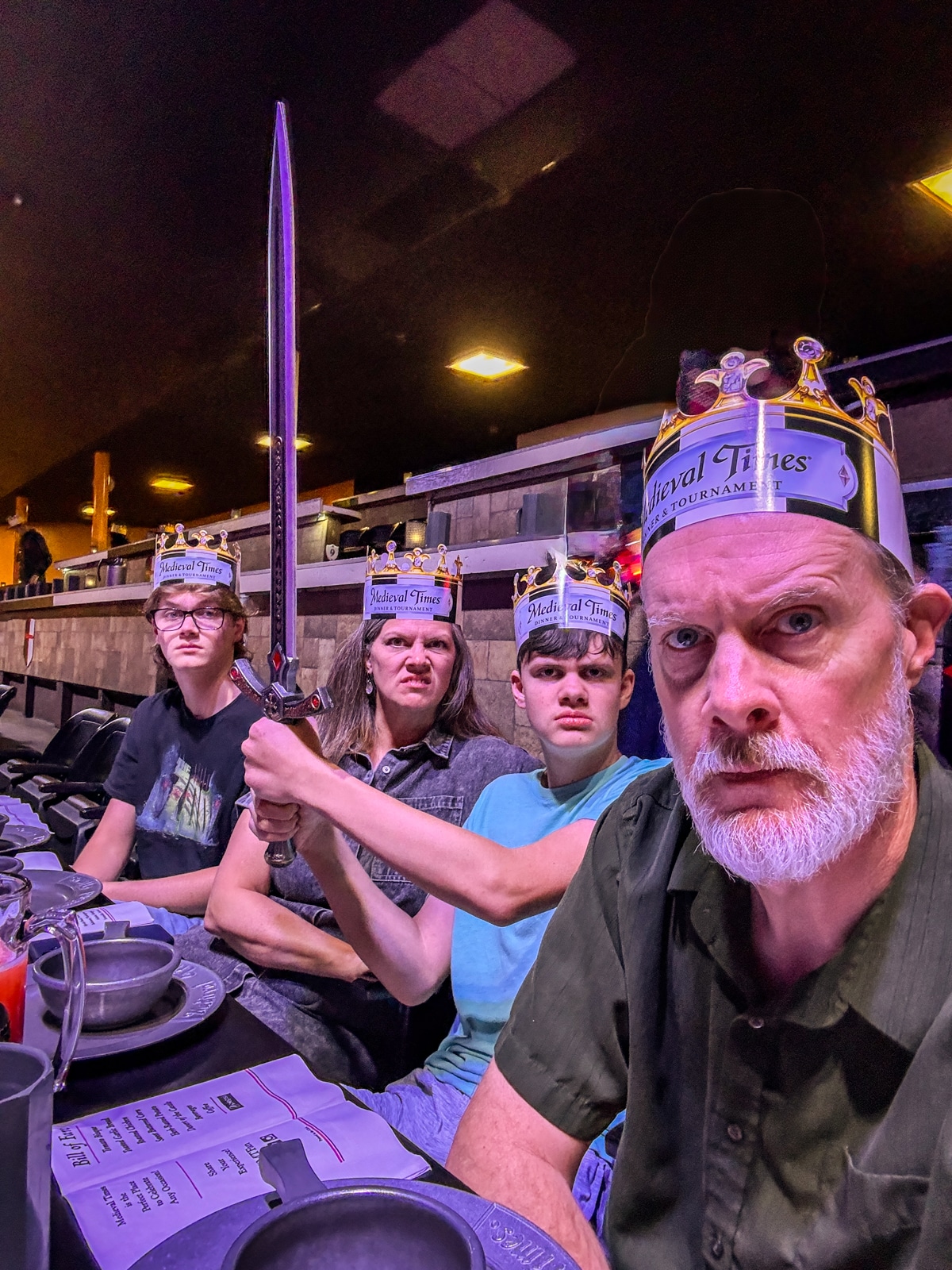 Four people wearing paper crowns sit at a dining table; one person holds a sword and makes a fierce face, while the others look at the camera with neutral or serious expressions.