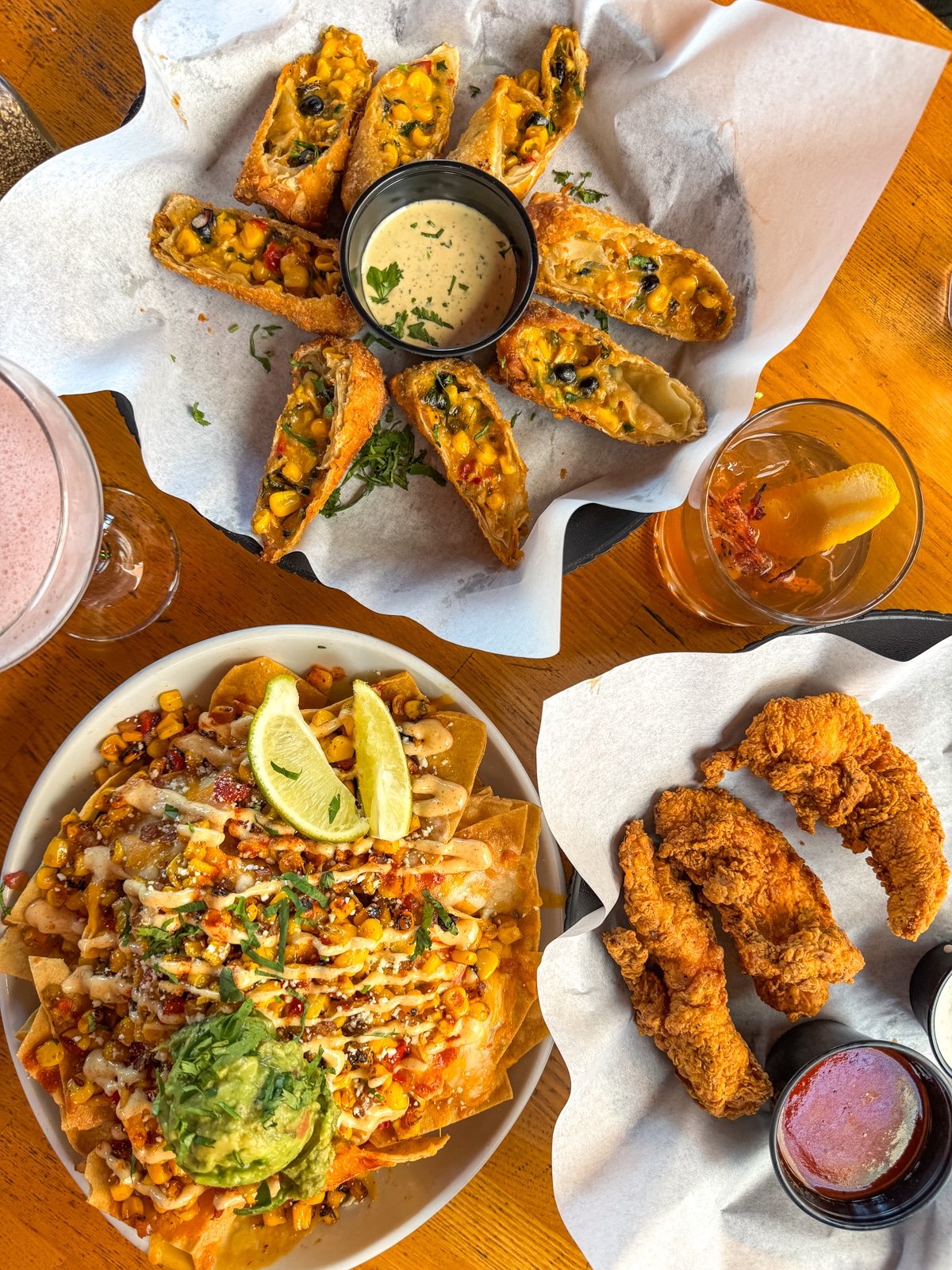 Three dishes on a wooden table: southwest egg rolls with dip, fried chicken tenders with sauce, and nachos topped with guacamole, corn, cheese, and lime wedges. Two drinks are nearby.