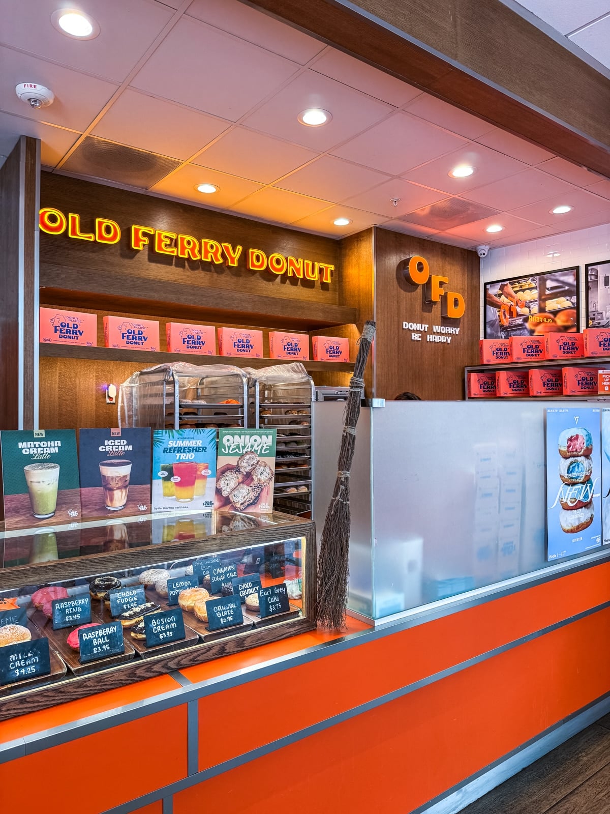 Counter at a donut shop named Old Ferry Donut, displaying various donuts and drinks, with orange boxes stacked on shelves and a broom leaning against the counter.
