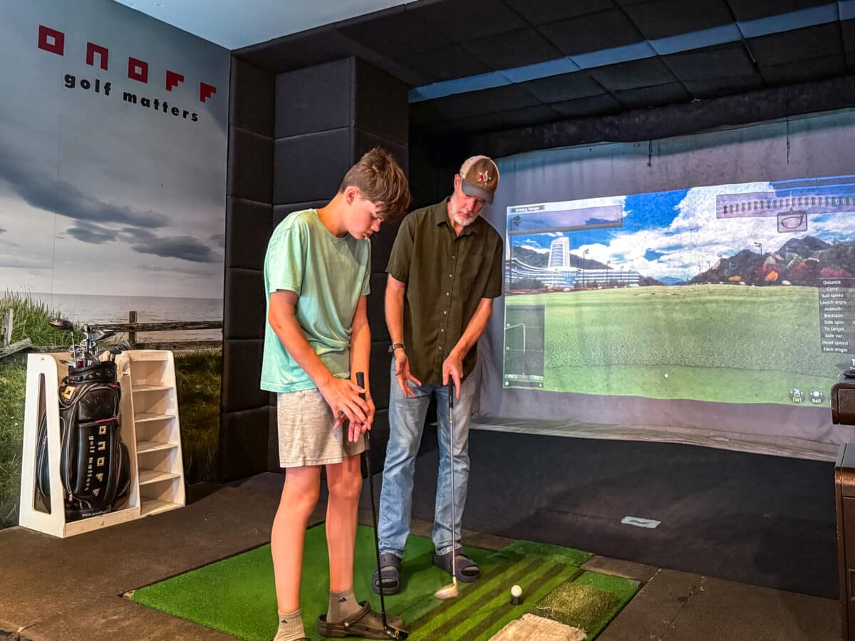 A man instructs a boy on how to putt at an indoor golf simulator, with golf clubs and a "golf matters" sign visible in the background.