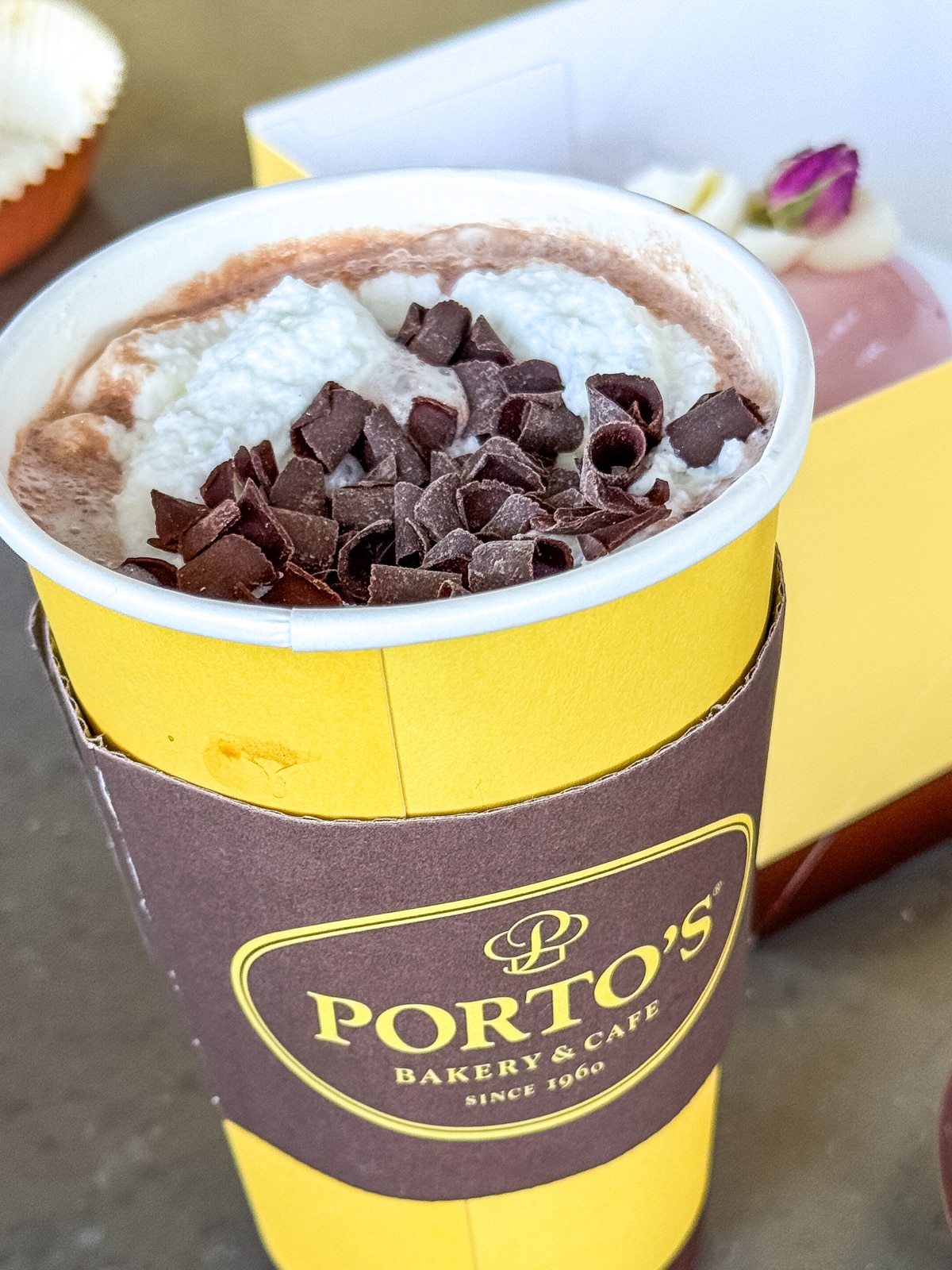 A cup of hot chocolate topped with whipped cream and chocolate shavings in a Porto's Bakery & Caf&eacute; cup, with pastries in the background.