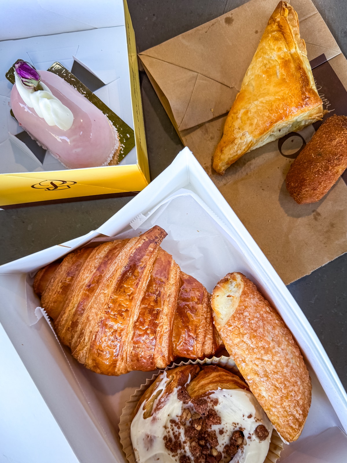 A selection of pastries including a pink mousse cake, a turnover, a croquette, a croissant, a danish, and a cinnamon roll, displayed in boxes and on paper.