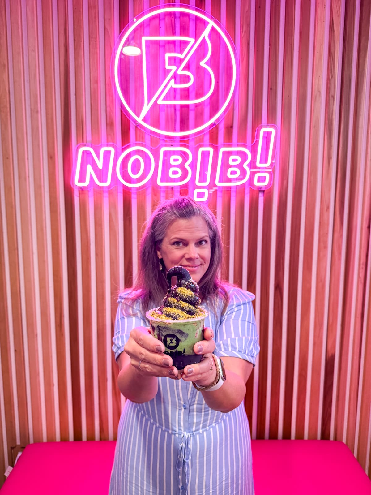 A woman in a blue striped dress holds a cup of colorful soft serve ice cream in front of a neon "NOBIBI" sign and wooden wall.