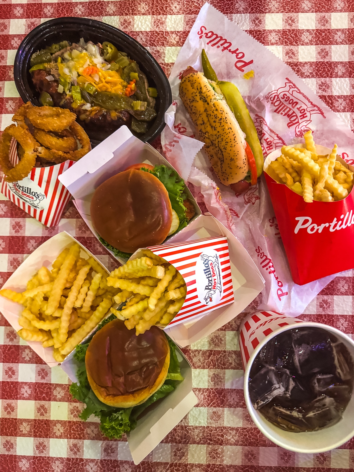 A table with two burgers, crinkle-cut fries, onion rings, a chili cheese dog, a cup of soda, and a container of fries on a red and white checkered tablecloth.