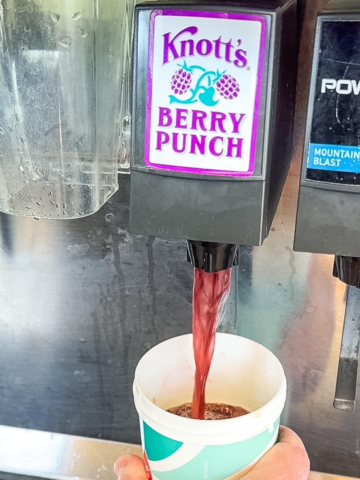 A beverage dispenser pours Knott's Berry Punch into a paper cup held by a person.