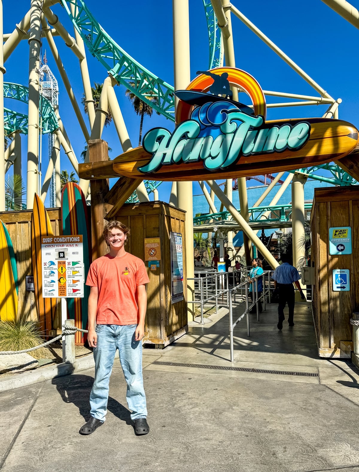 A person stands smiling in front of the HangTime roller coaster entrance at an amusement park, with surfboards and a ride sign visible.