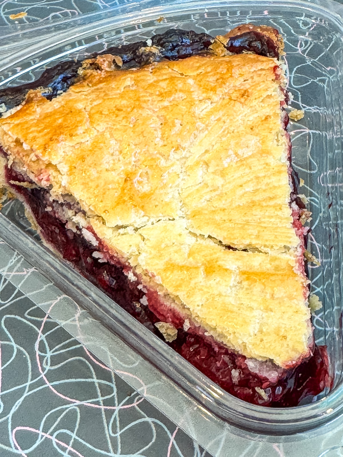 A single slice of fruit pie with a golden, flaky crust and dark berry filling sits in a clear plastic container.