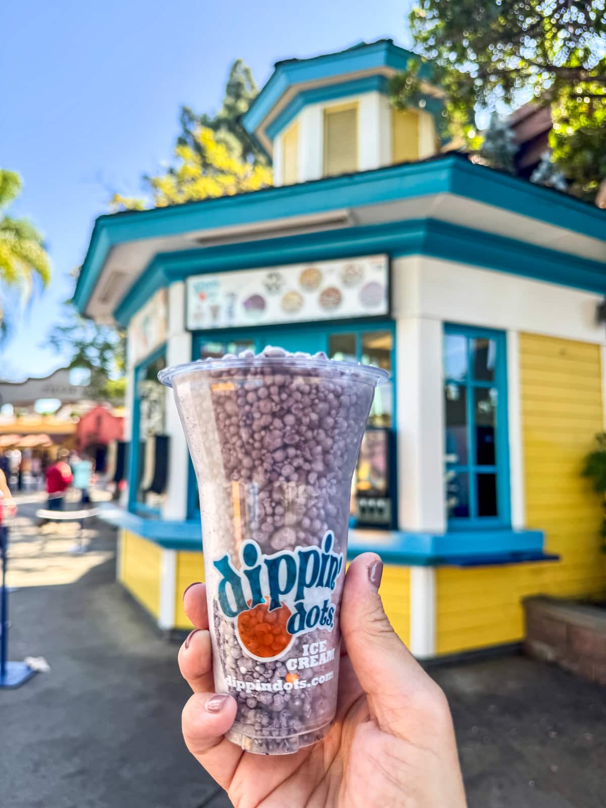 A hand holds a cup of Dippin' Dots ice cream in front of a colorful kiosk with a blue and yellow exterior.