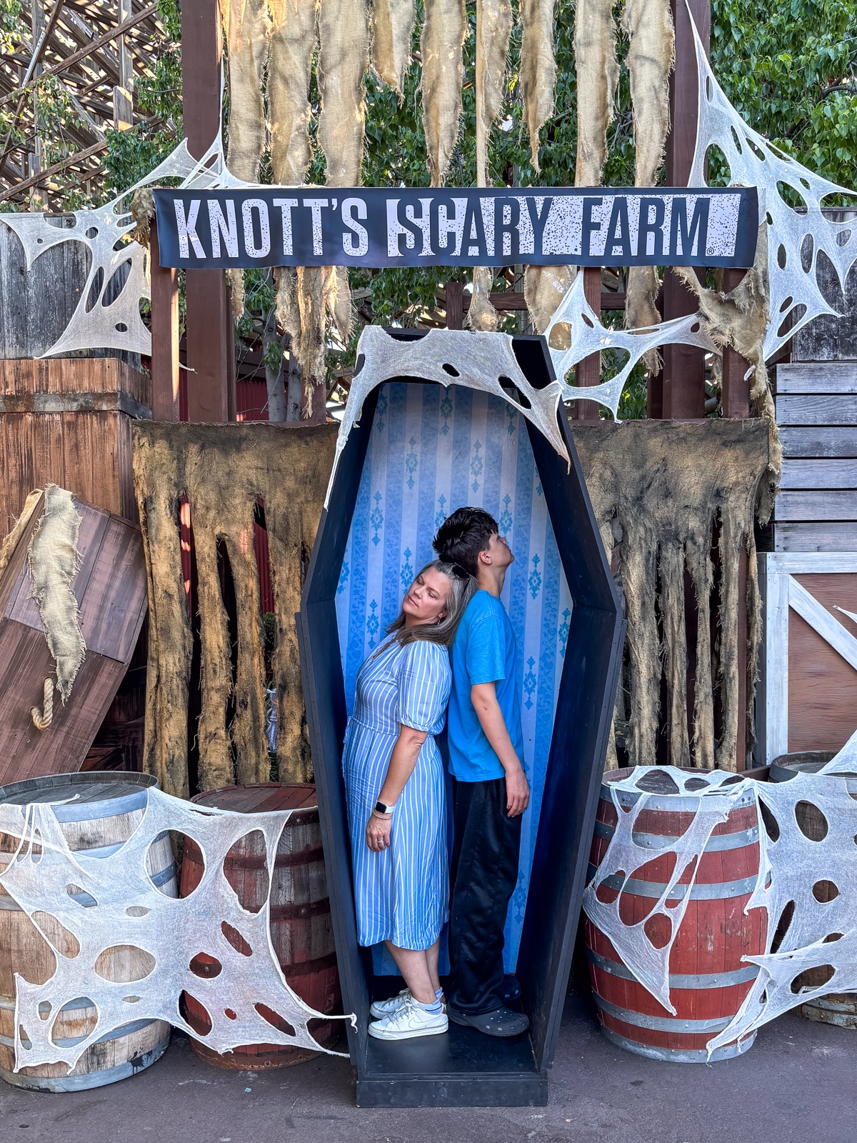 Two people stand back-to-back inside a coffin prop at Knott's Scary Farm, surrounded by Halloween decorations and faux cobwebs.