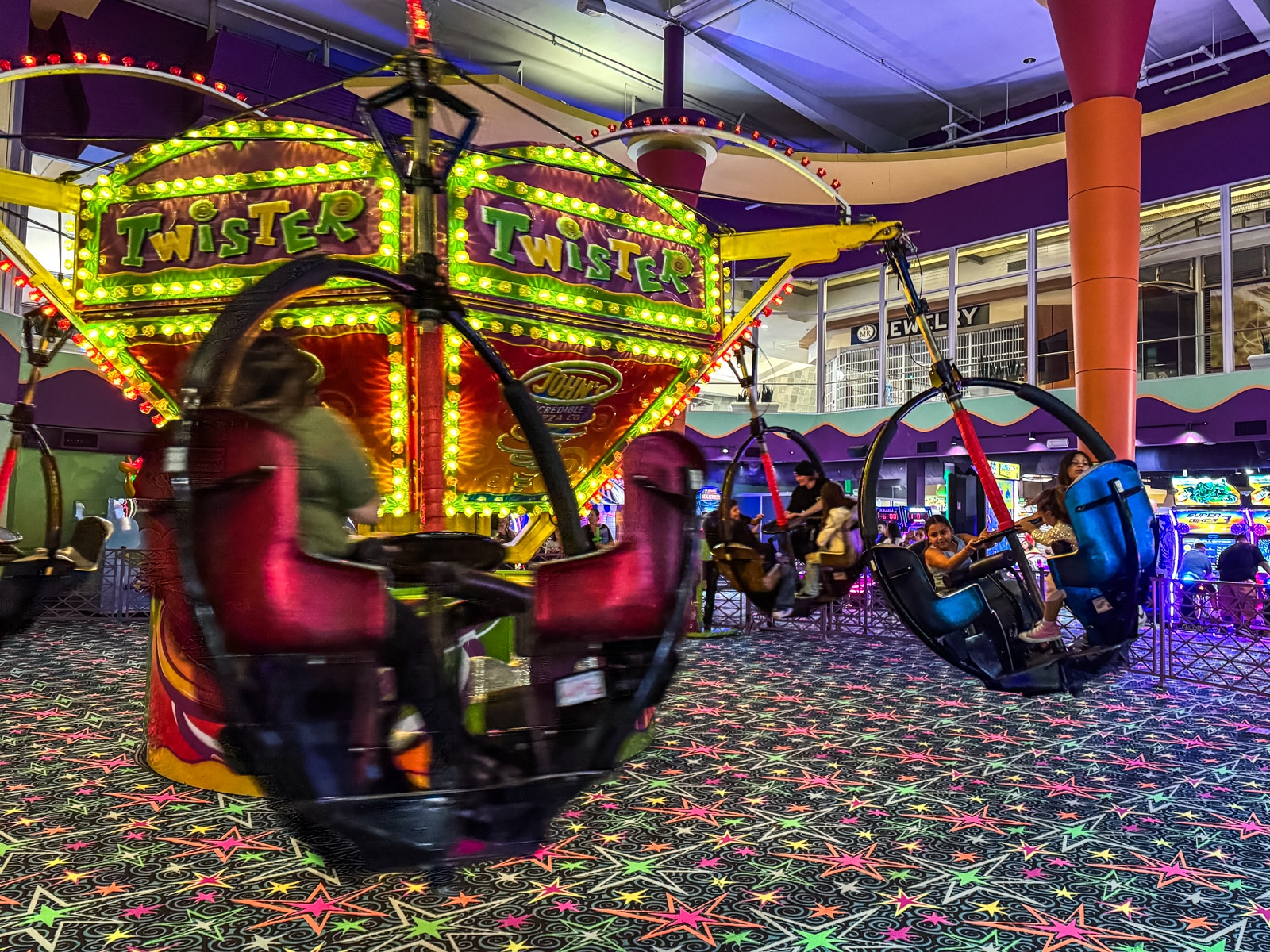 A spinning amusement ride called Twister with children seated in hanging seats, surrounded by colorful lights and arcade games inside an indoor entertainment center.