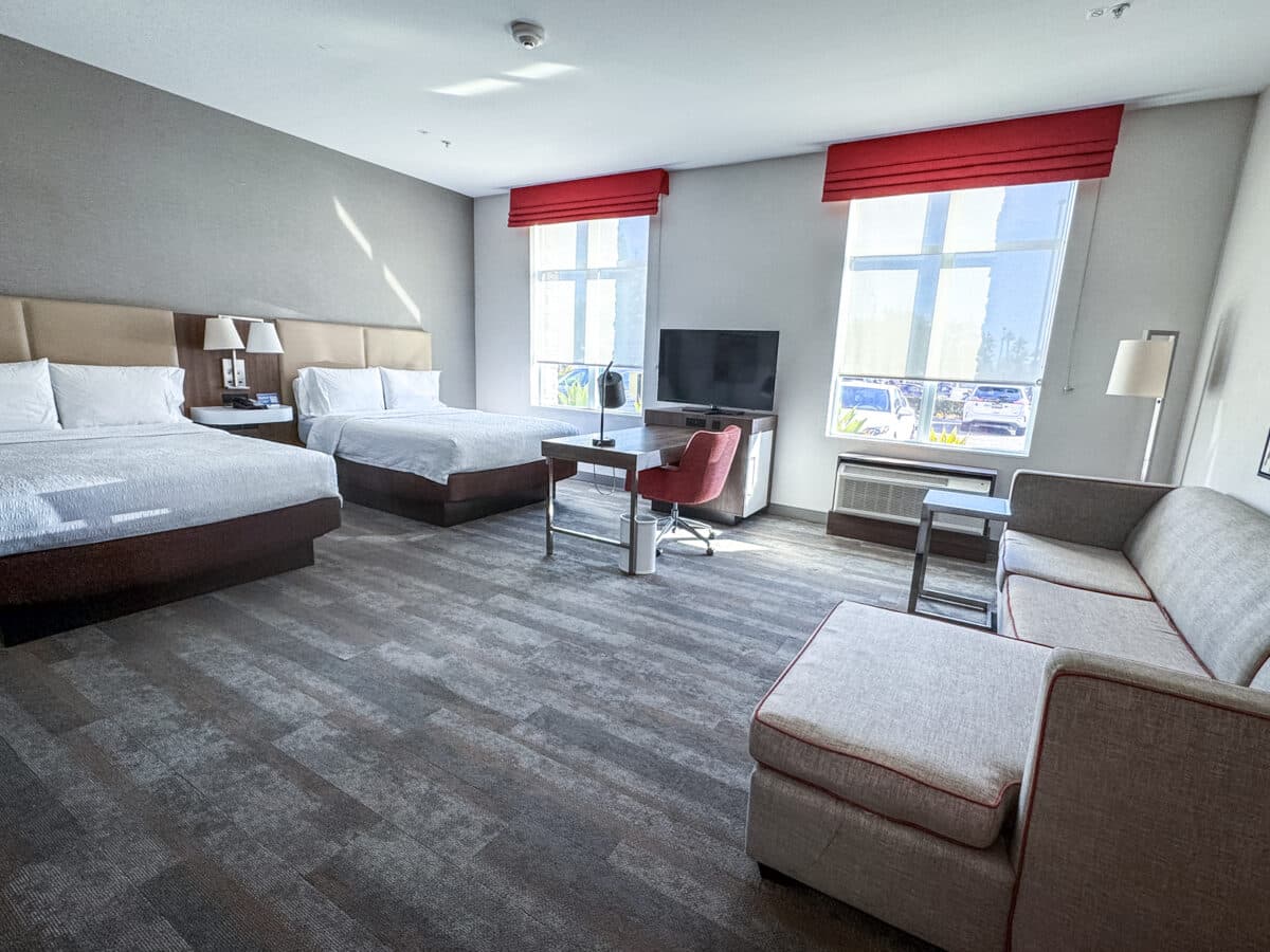 Hotel room with two queen beds, a couch, desk with chair, TV, lamps, and large windows letting in natural light.