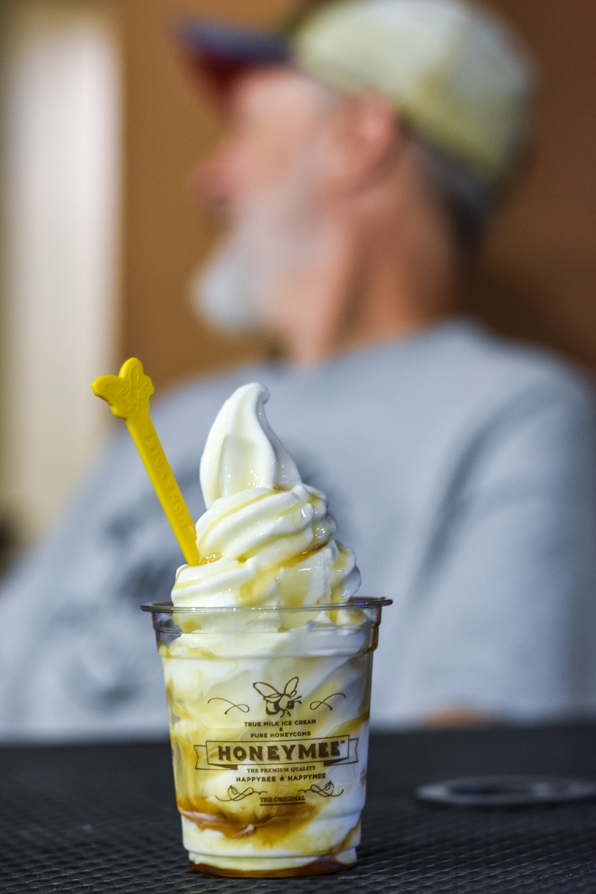 A close-up of a honey-drizzled soft serve ice cream in a cup with a yellow spoon, with a blurred person sitting in the background.