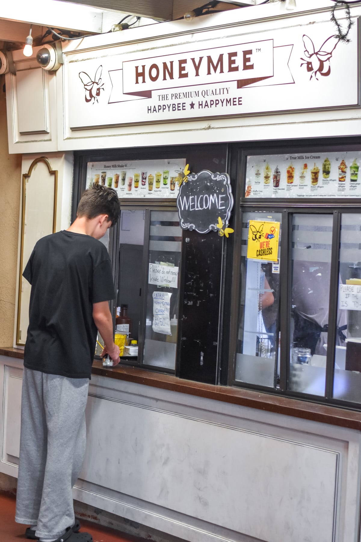 A person stands at the counter of a Honeymee dessert shop, looking down while preparing to order. The shop has a menu and welcome sign displayed above the window.