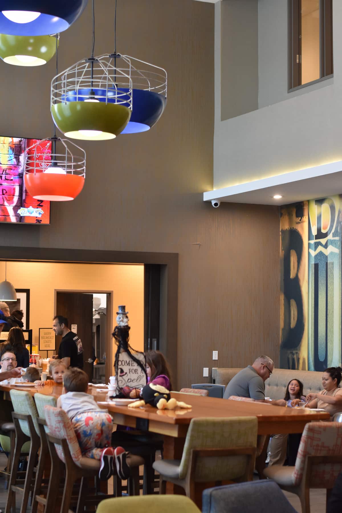 People sitting and talking at tables in a modern restaurant with colorful hanging lights and a large mural on the wall.