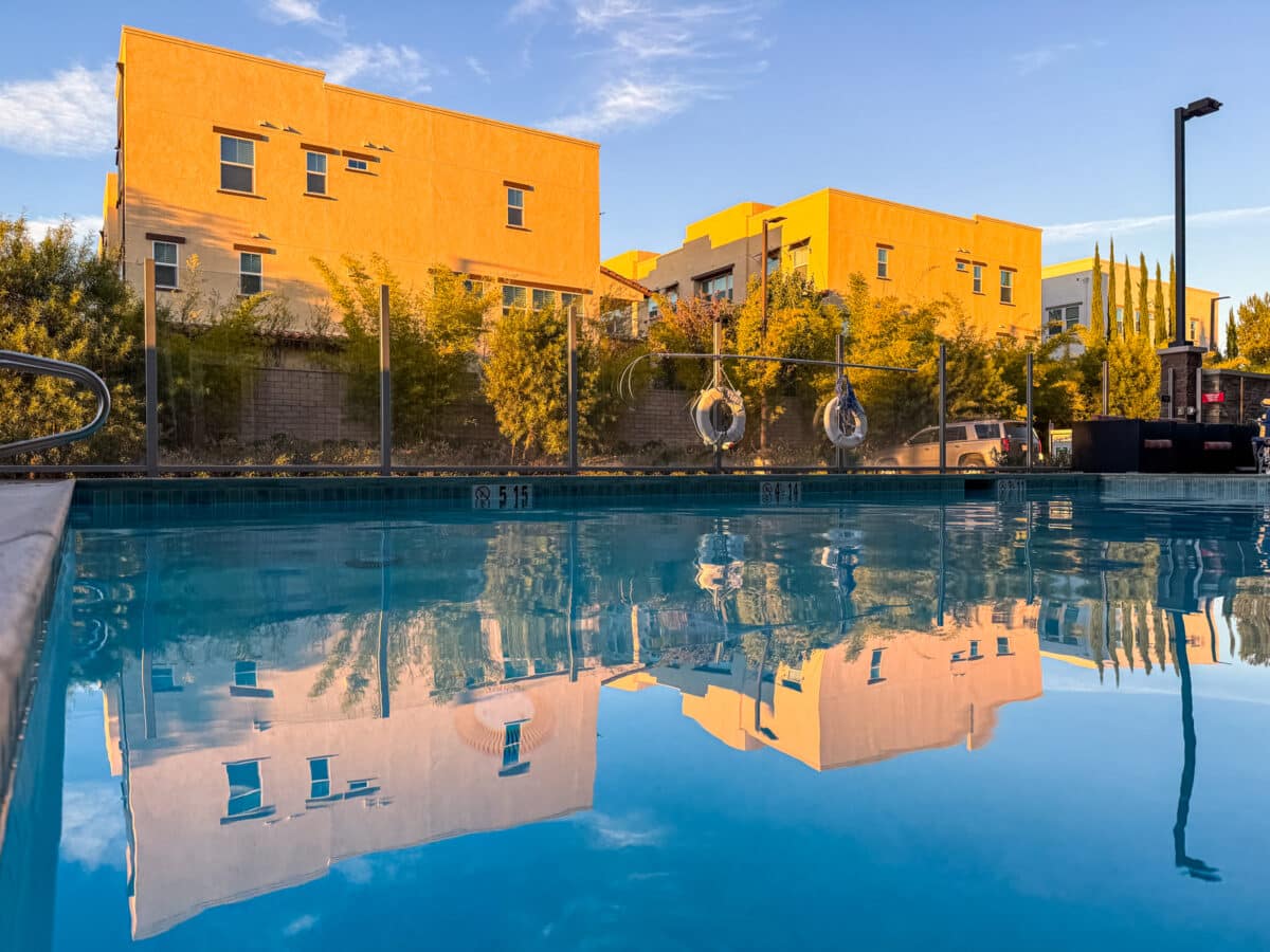Apartment buildings and trees are reflected in a clear outdoor swimming pool, with blue sky and sunlight illuminating the scene.