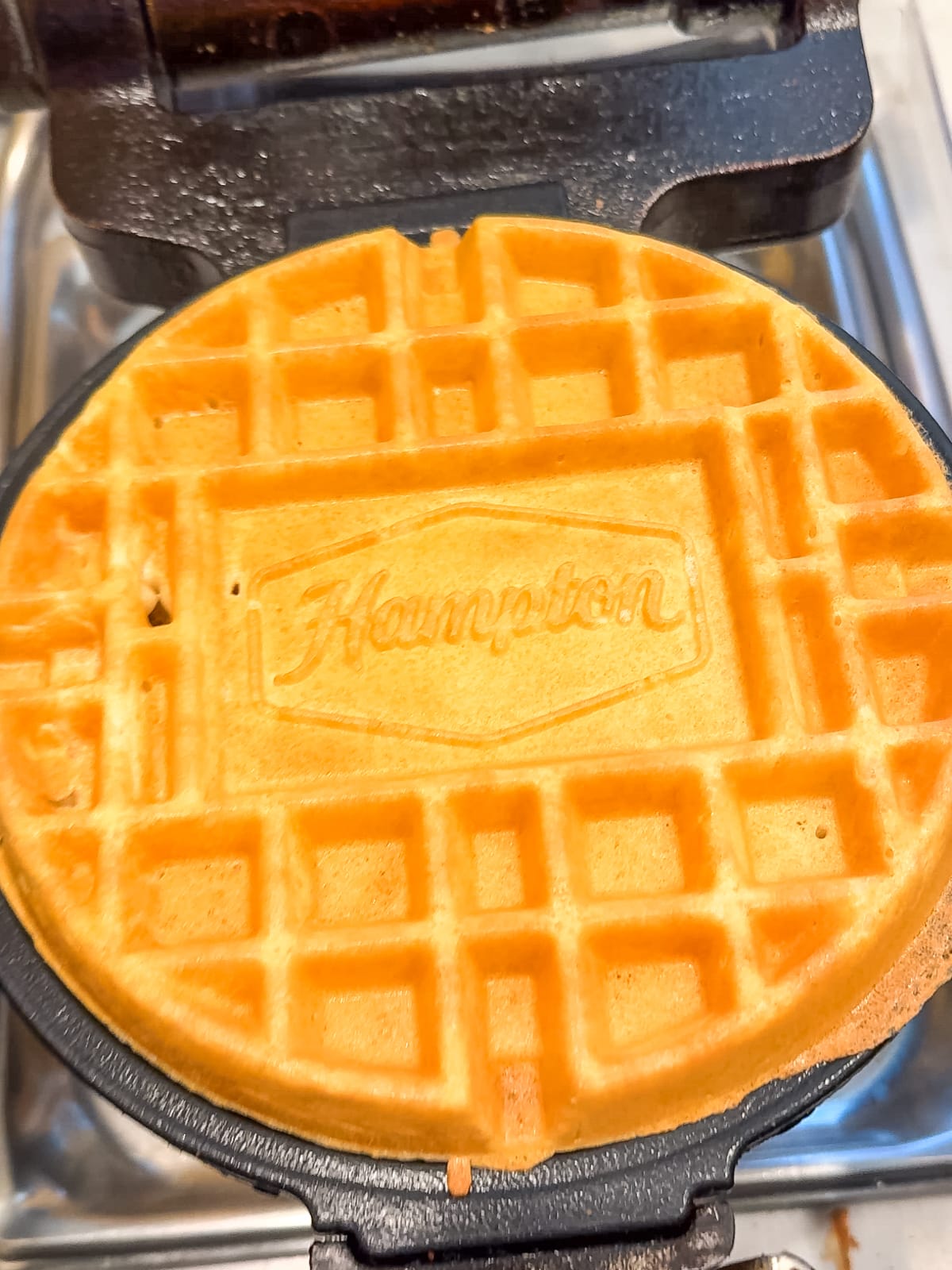 A golden waffle with the word "Hampton" imprinted on it sits on a waffle maker.