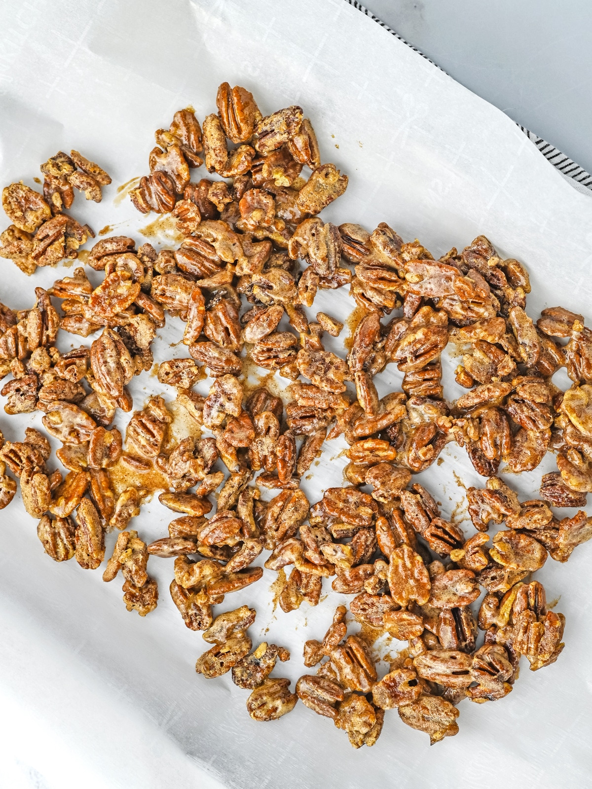 Pecans coated in a glossy mixture are spread out on a sheet of parchment paper on a baking tray.