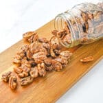A glass jar tipped over on a wooden board with candied pecans spilling out onto the surface.