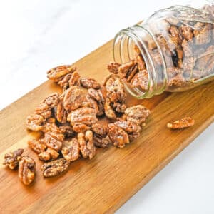 A glass jar tipped over on a wooden board with candied pecans spilling out onto the surface.