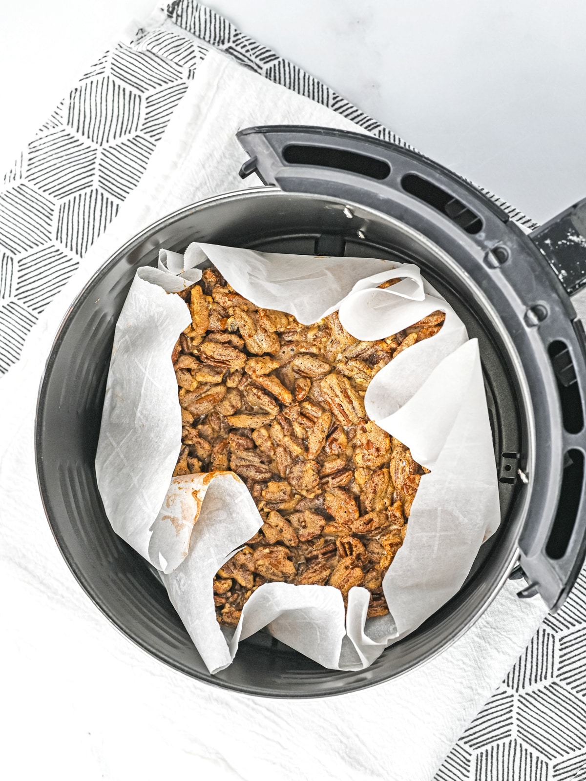 A baked casserole topped with crispy pieces sits on parchment paper inside an air fryer basket, placed on a patterned cloth.