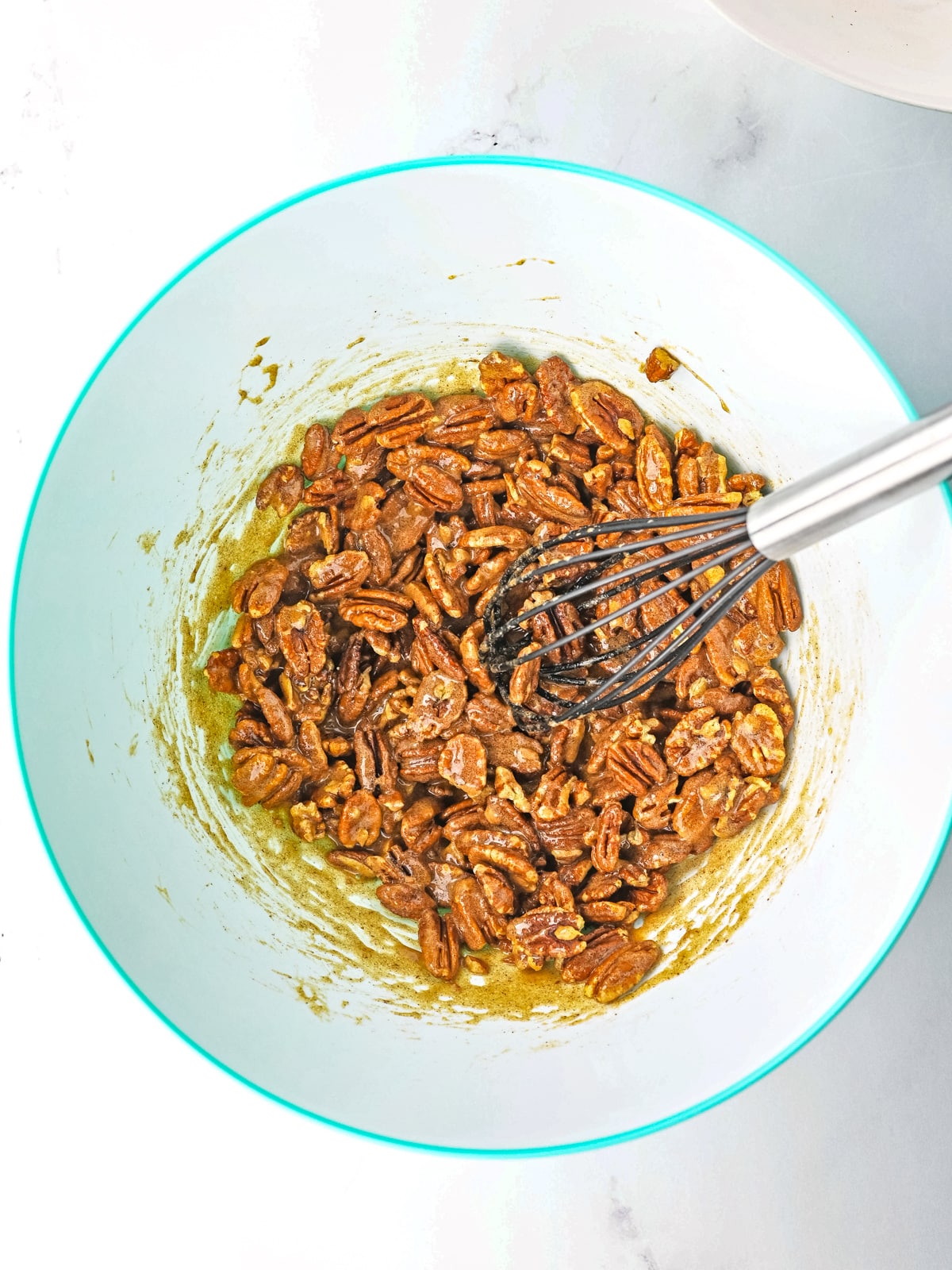 A metal whisk mixes pecans with a brown sugary coating in a large light blue mixing bowl on a white surface.