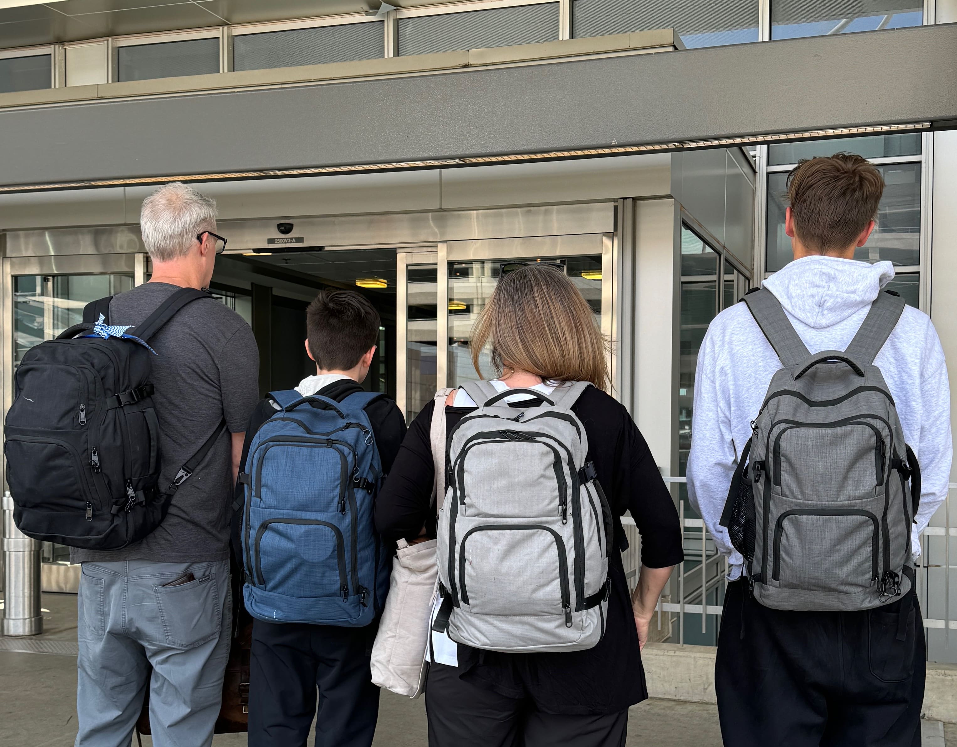 Four people with backpacks stand in front of sliding glass doors at an entrance, facing away from the camera.