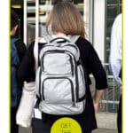 A woman with a gray backpack enters a building through glass doors, with two other people walking nearby. Text overlay reads "Souvenir Survival Kit" and "Get the List.